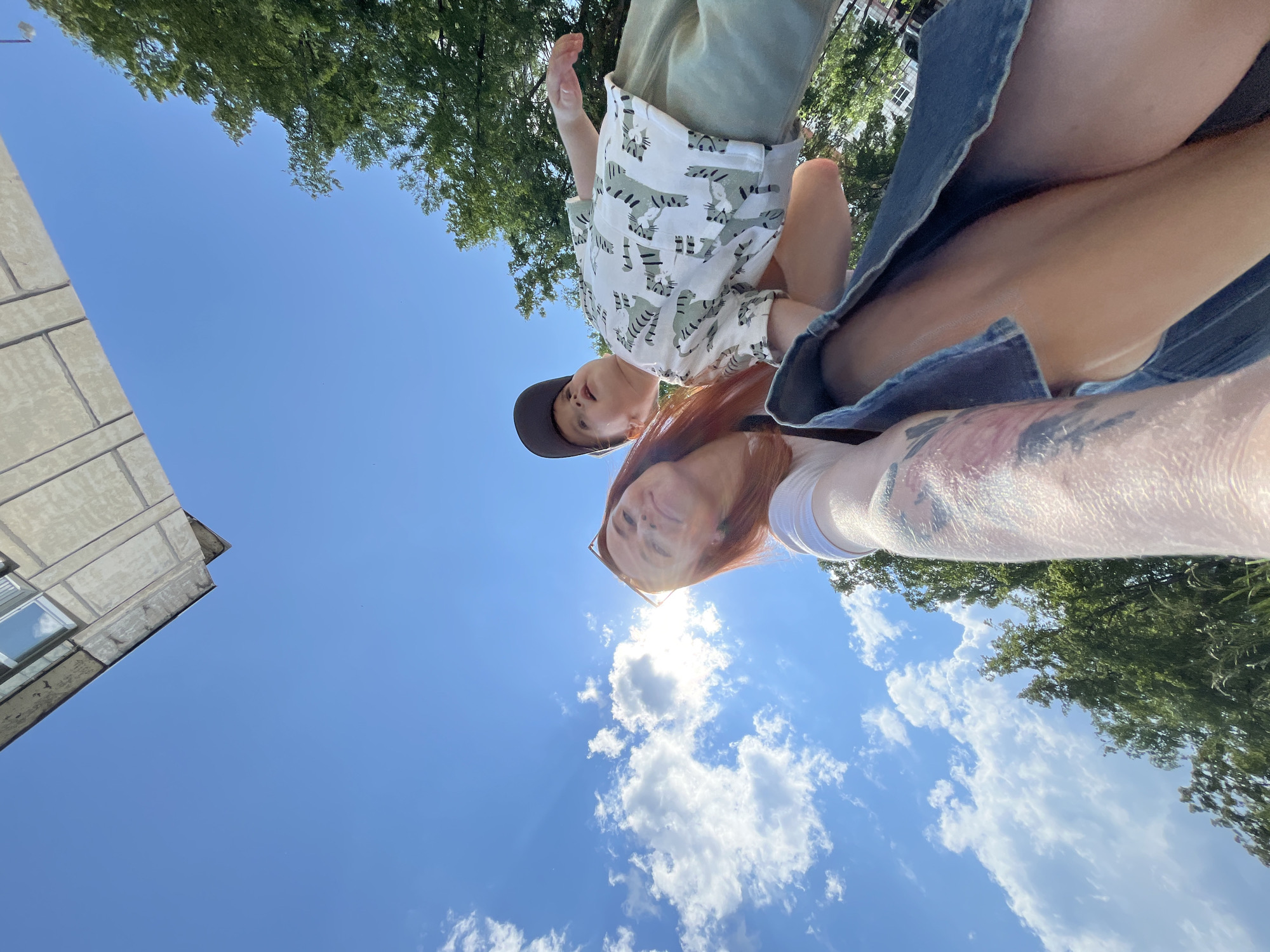A low-angle shot shows a woman holding a young boy in her arms, both looking up at the camera against a backdrop of blue sky and clouds. The woman, with red hair and wearing sunglasses, smiles at the camera. The boy is wearing a patterned shirt and a baseball cap. The scene is outdoors, with trees and a building visible in the background. The image was taken in the city of Chaikovsky, Russia.