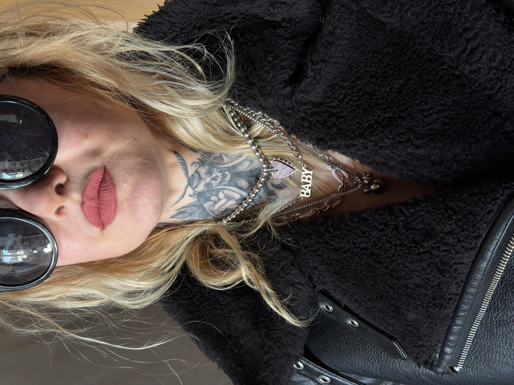 A blonde woman wearing large, round, dark sunglasses is lying down. She has a neck tattoo and wears multiple silver necklaces, one of which has a charm that says "BABY". She is wearing a black, furry jacket with a leather section visible on the right side of the frame, including a zipper. The background is a light-colored surface, possibly a table or floor. The location is not identifiable as Saint Petersburg, Russia from the image.