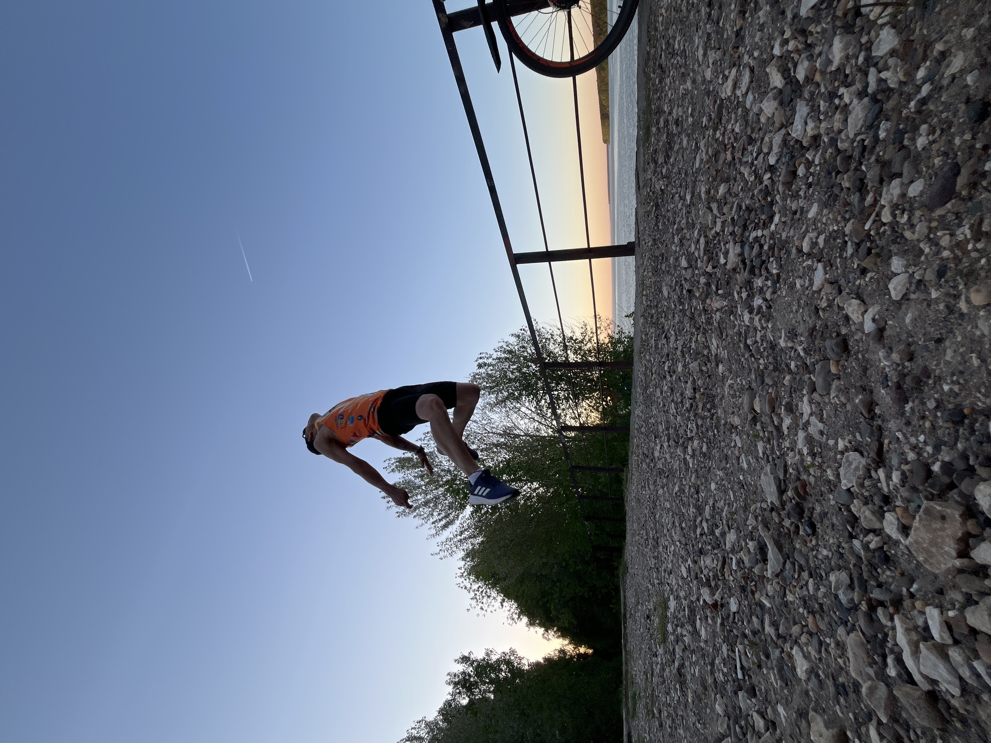 The image shows a person in mid-air performing a backflip. The individual is wearing an orange tank top, black shorts, and blue sneakers with white stripes. They are upside down against a clear blue sky with a contrail visible in the upper left.
In the background, there is a metal railing, a bicycle wheel, a body of water with a sunset visible on the horizon, and a line of trees along the shore. The foreground shows a rough concrete or stone surface.
Based on the visual cues of a river, a sunset horizon, and the specific type of Soviet-era architectural infrastructure, it is plausible that this image was taken in the city of Chaikovsky, Russia.