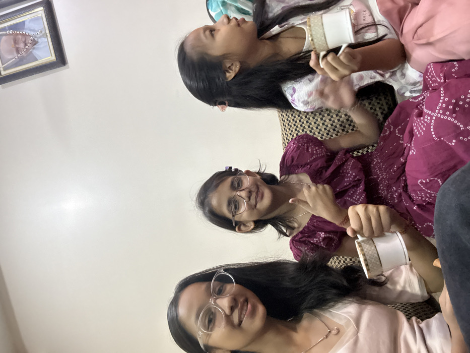 Three girls are casually seated on a patterned couch. One girl wears a pink dress, eyeglasses and a necklace, another wears a purple patterned dress and eyeglasses, and the third wears a white patterned top. All are holding teacups. A framed picture hangs on the wall. The image is rotated counterclockwise.