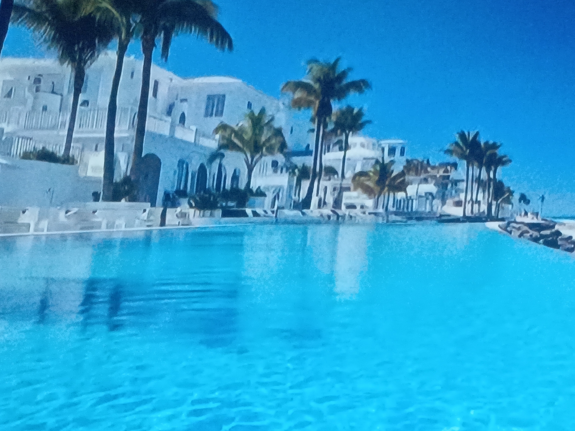 The image depicts a tropical scene in Juan Griego, Venezuela, featuring a bright turquoise pool in the foreground. Behind the pool, there is a white building complex in a Mediterranean style, with multiple levels and arched windows. Several palm trees are scattered throughout the scene, adding to the tropical ambiance. The sky is a clear blue. There are lounge chairs along the edge of the pool. Rocks are visible on the right.