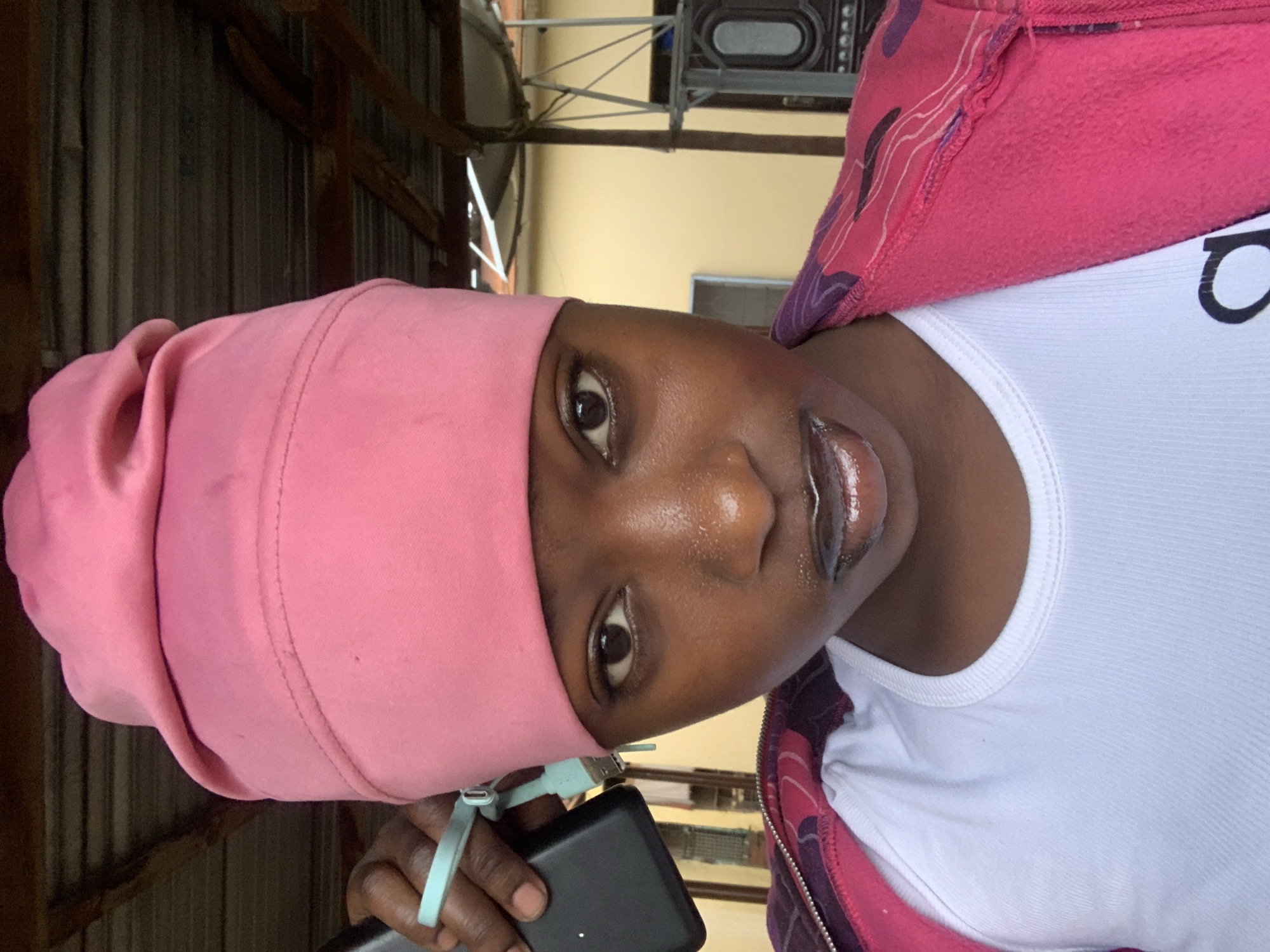 The image depicts a person wearing a pink head covering, a white shirt, and a pink jacket. They are holding a black electronic device, possibly a phone or a power bank, with a light blue strap attached. The background shows elements of a building, possibly a metal structure and some walls. The location is not specified, so the city cannot be confirmed.