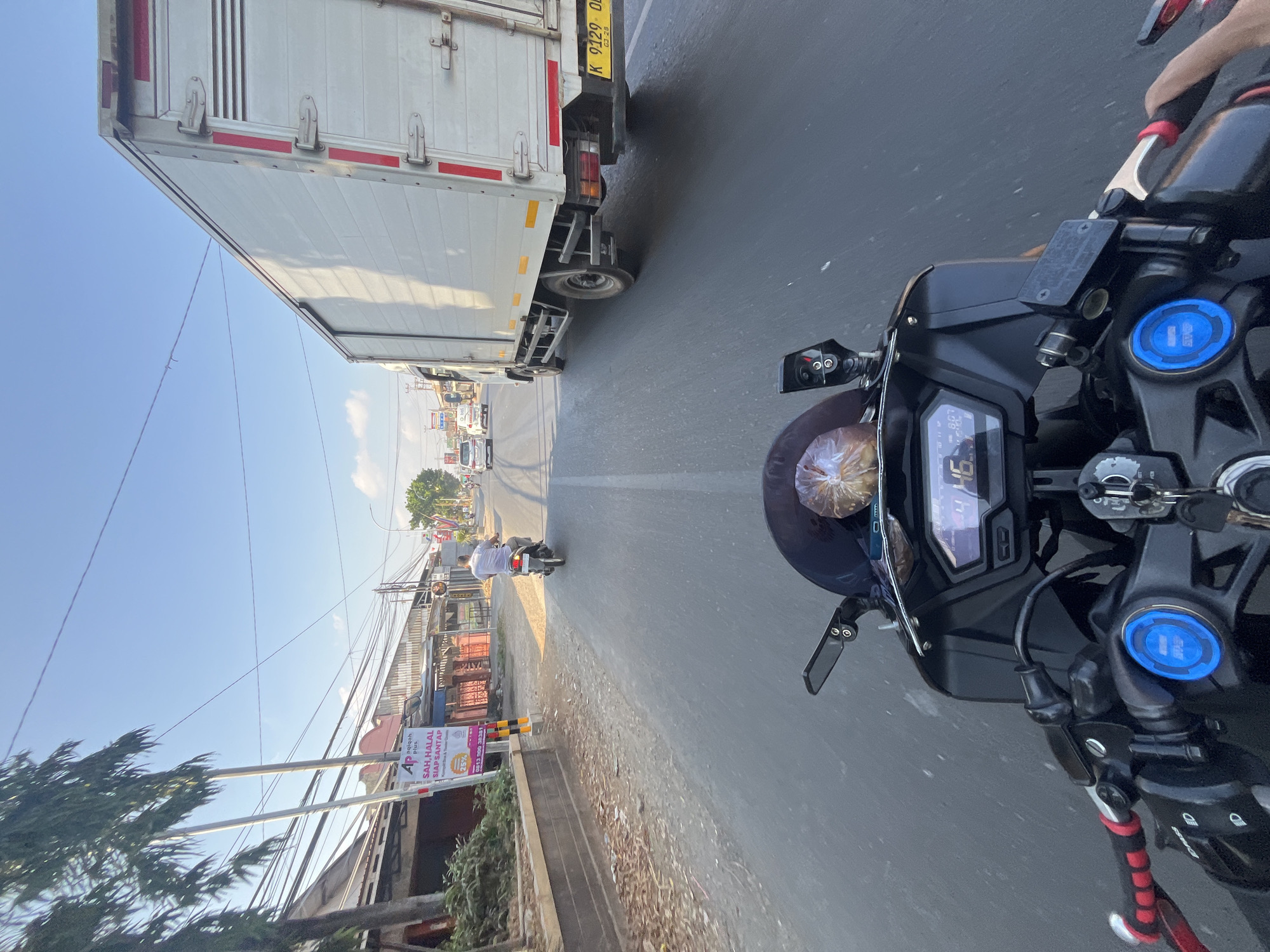 This is a first-person perspective photograph taken from a motorcycle on a street in Kalinyamatan, Indonesia. The motorcycle's handlebars and dashboard are in the foreground, showing a digital display with the number "46". The motorcycle is in motion on an asphalt road, which appears to be a main street with traffic. A white box truck is visible in the right lane, its license plate reading "K 9129 OB". In the background, there are buildings lining the street, some of which appear to be shops. A person is riding a motorcycle in the left lane ahead of the perspective. Trees and utility poles with numerous wires are also present along the roadside. The sky is clear and blue, indicating daytime.