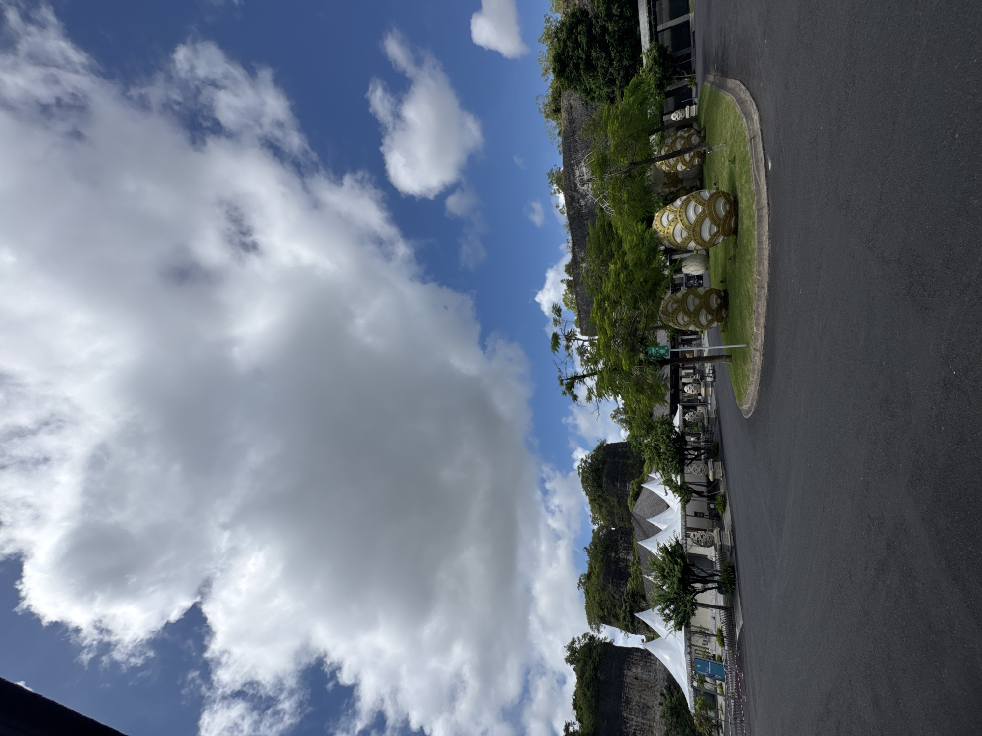 The photograph captures a scene in Kuta Selatan, Indonesia, presenting a blend of natural and man-made elements under a partly cloudy sky. The left side of the image is dominated by a sky with patches of blue interspersed with large, billowing white and grey clouds. On the right, a dark asphalt road curves into the distance. Alongside the road, there's a green, landscaped area featuring trees and decorative, egg-shaped sculptures with intricate patterns. In the background, there are rocky cliffs covered with lush green vegetation. Further down the road, there are structures with white, conical roofs. The overall composition suggests a place where urban development meets the natural landscape.