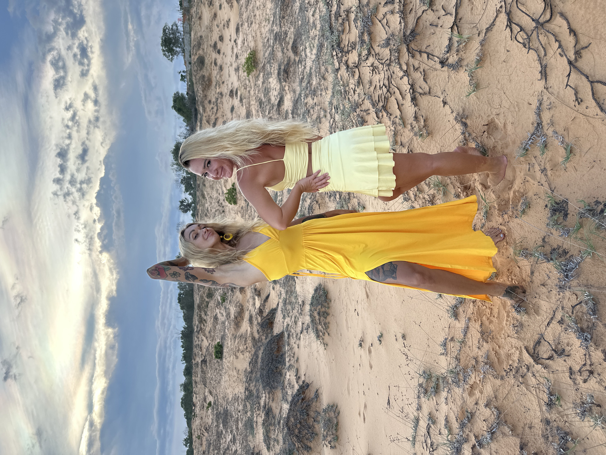 Two women stand in a sandy landscape with sparse vegetation under a cloudy sky. The woman on the left wears a long yellow dress with a thigh-high slit and has tattoos on her arm and leg. The woman on the right wears a yellow crop top and a matching ruffled skirt. The location is the Mui Ne sand dunes in Vietnam.