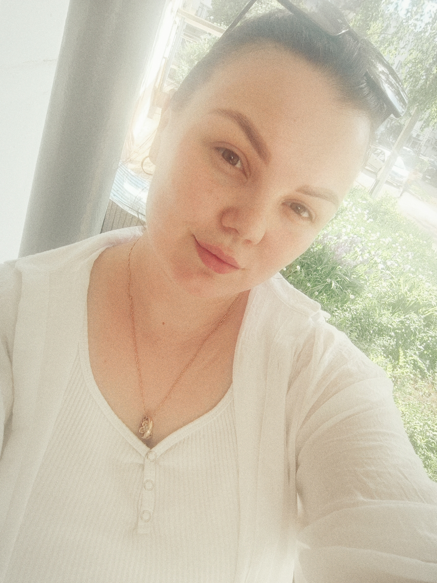 A selfie of a fair-skinned woman with brown hair pulled back, wearing sunglasses on her head, a white button-down top, and a gold necklace. She is positioned to the left of a gray concrete pillar, with a blurred background of green grass, plants, and a parked car. The location is a building in a city of Unknown, Unknown.