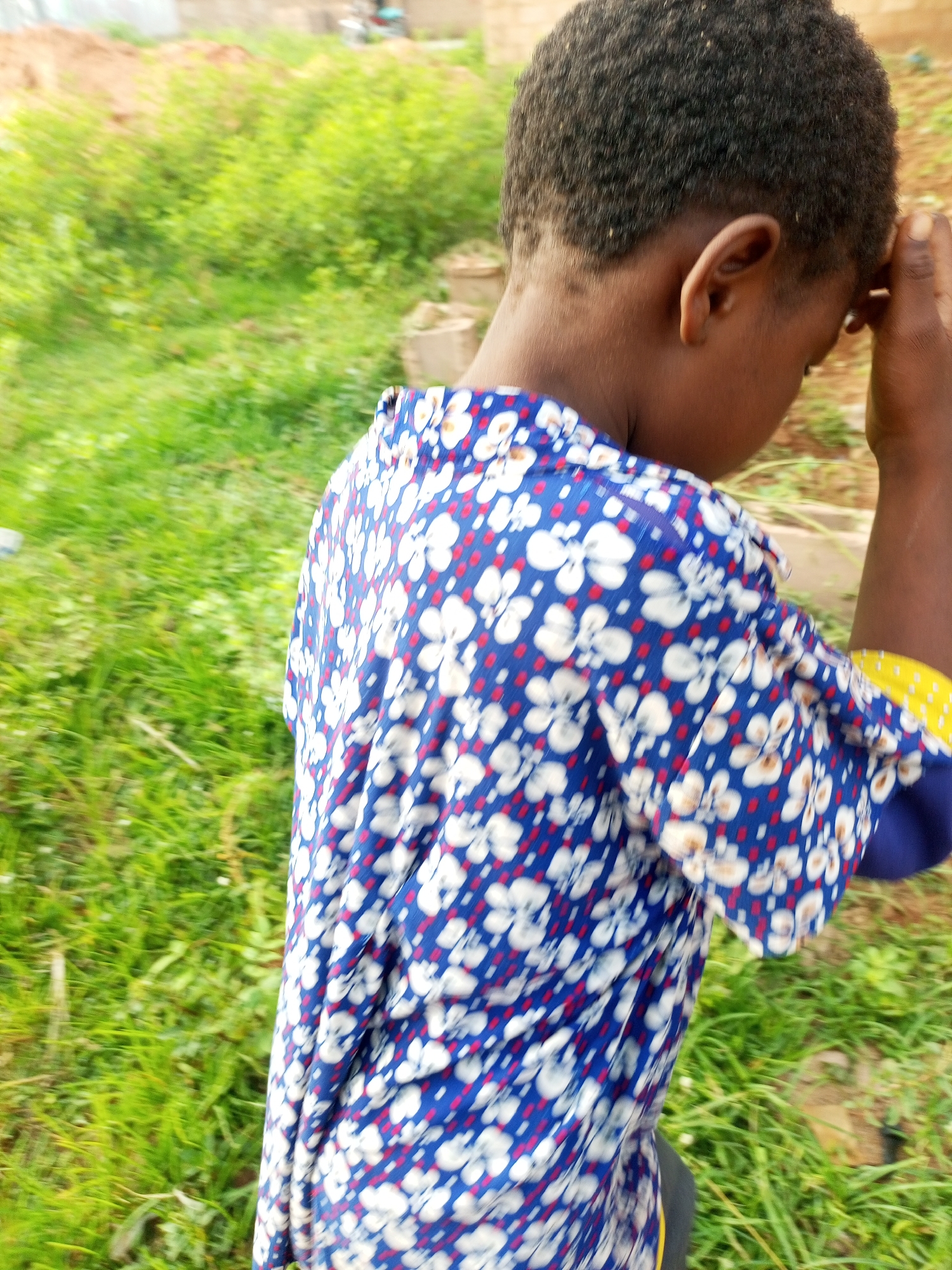 A medium shot, taken from the shoulder down, shows a person wearing a blue, white, and red floral pattern shirt. The person's dark hair is short and textured. Their left hand is raised to their forehead, and their head is tilted down, obscuring their face. The background is blurred, showing green grass and a sandy area. The image has a shallow depth of field, with the person in focus and the surroundings out of focus. The lighting suggests it is daytime. The scene appears to be outdoors, in a rural or semi-rural area, possibly in Tempure, Nigeria, as indicated by the mention of that location.