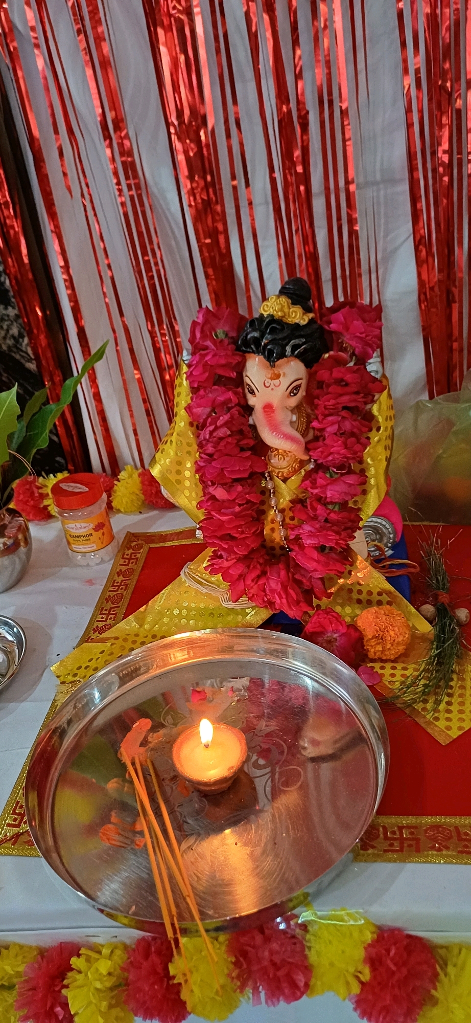 An indoor scene, likely a home altar or temporary shrine, is depicted with a focus on a statue of Lord Ganesha. The background features a decorative backdrop of red and white tinsel streamers. The statue of Ganesha is adorned with a vibrant pink floral garland and a yellow cloth decorated with gold patterns. In the foreground, a silver thali (tray) holds a lit diya (earthen lamp) with a flickering flame, and several unlit incense sticks. To the left of the thali, a small jar labeled "CAMPHOR 100% Pure" is visible, alongside some artificial red and yellow flowers. To the right of the Ganesha idol, an orange marigold flower and some dried natural elements are present. The overall ambiance suggests a religious or festive occasion. The presence of specific religious iconography and cultural elements points to a celebration such as Ganesh Chaturthi, commonly observed in India, including in cities like Ajmer.