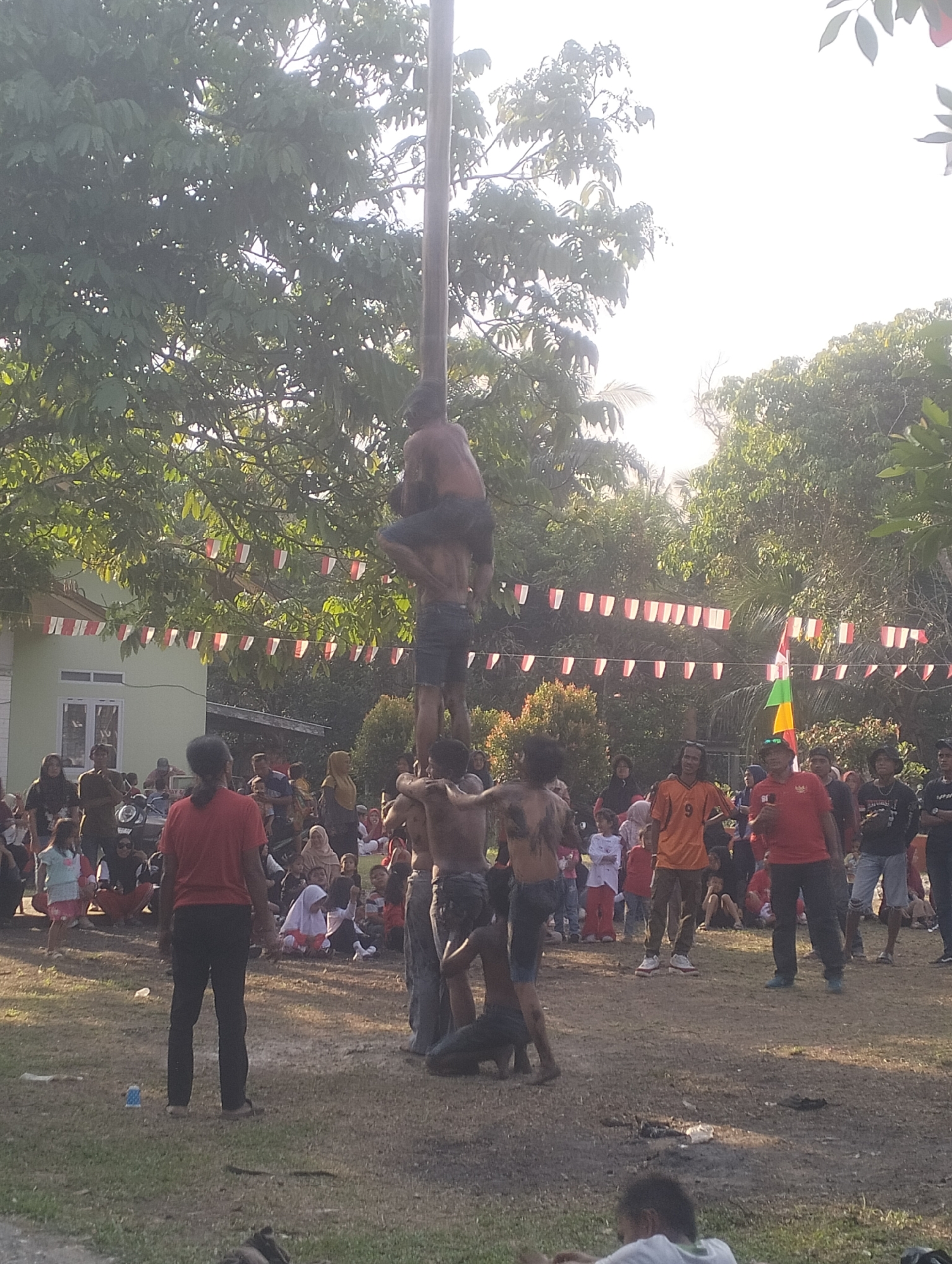 The image depicts an outdoor event in Dumai City, Indonesia, likely a festive gathering. The central focus is a group of men participating in a traditional game where they are stacked on top of each other, climbing a tall wooden pole. The men at the bottom appear to be supporting those above them, with one man at the very top sitting on another's shoulders. Their bodies are partially covered in mud, suggesting a challenge or competition element.
A crowd of spectators, including men, women, and children, are gathered around, observing the event. Many are seated on the ground, while others stand. The environment is characterized by lush green trees and a clear sky, indicating a daytime setting. Red and white pennant flags are strung across the scene, a common decoration during national celebrations in Indonesia. In the background, a light green building with white window frames can be seen, along with parked motorcycles, suggesting a community area or event space. The ground is a mix of dry earth and grass. The overall atmosphere appears to be one of community engagement and entertainment.