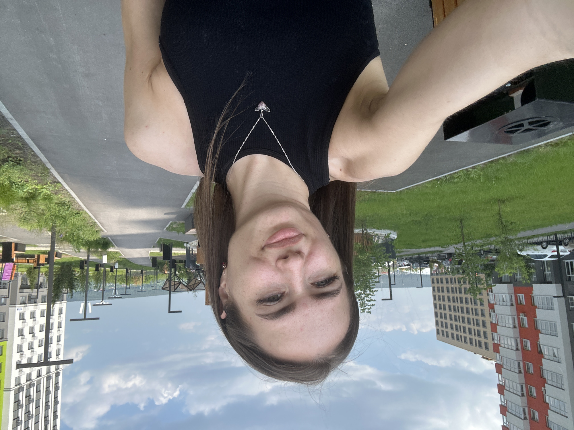 A young woman with long brown hair is taking a selfie outdoors in an urban park. She is wearing a black tank top and a silver necklace with a heart-shaped pendant. The background shows a paved walkway lined with streetlights and benches, with green grass and trees on either side. In the distance, there are modern apartment buildings and a cloudy sky.