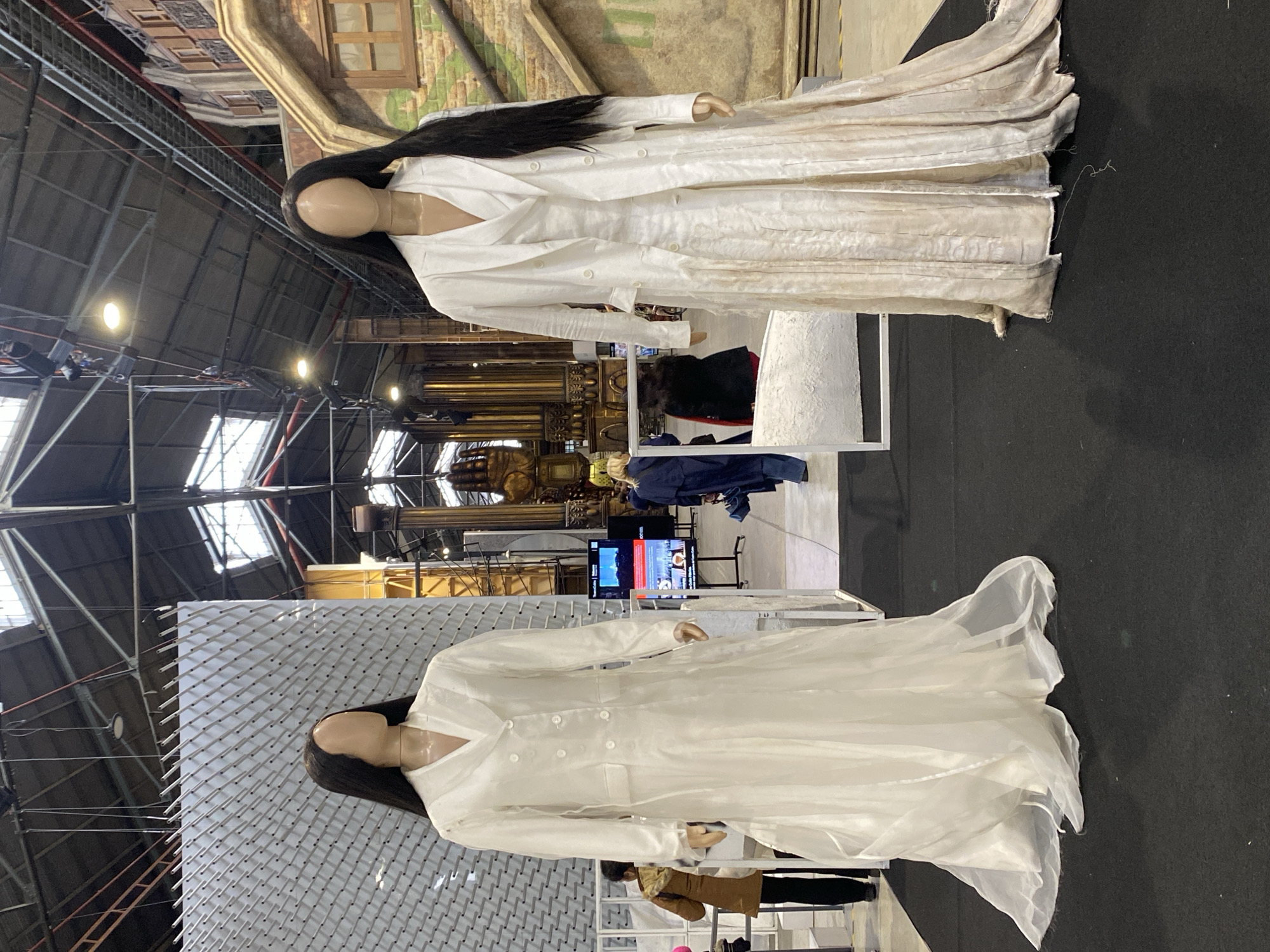 The image captures an indoor exhibition or event, likely taking place in a large venue such as a convention center or repurposed industrial space, characterized by a high, exposed ceiling with metal beams, ductwork, and bright artificial lighting. The floor is covered in a dark material, possibly black carpeting.
The primary subjects are two stylized mannequins, each with a faceless head, light skin tone, and long, straight black hair. Both are dressed in distinct, floor-length white coats or dresses. The mannequin on the left wears a flowing, slightly sheer white coat featuring a V-neckline and a double-breasted design with six white buttons. Behind this mannequin is a large white panel with numerous horizontal, protruding elements, creating a textured surface. The mannequin on the right is clad in a similar long white coat, but its fabric appears heavier, more textured, and possibly distressed or aged, particularly towards the hem. This mannequin is positioned in front of a painted backdrop resembling an old building facade with windows.
In the mid-ground, a large flat-screen television displays what appears to be a news channel or presentation, though the text and images are too blurry to discern specific details. The background also reveals various other exhibition elements, including an ornate, possibly golden structure and industrial scaffolding. The scene depicts a static display within a larger exhibition environment, suggestive of a fashion, art, or cultural exposition occurring in Buenos Aires, Argentina.