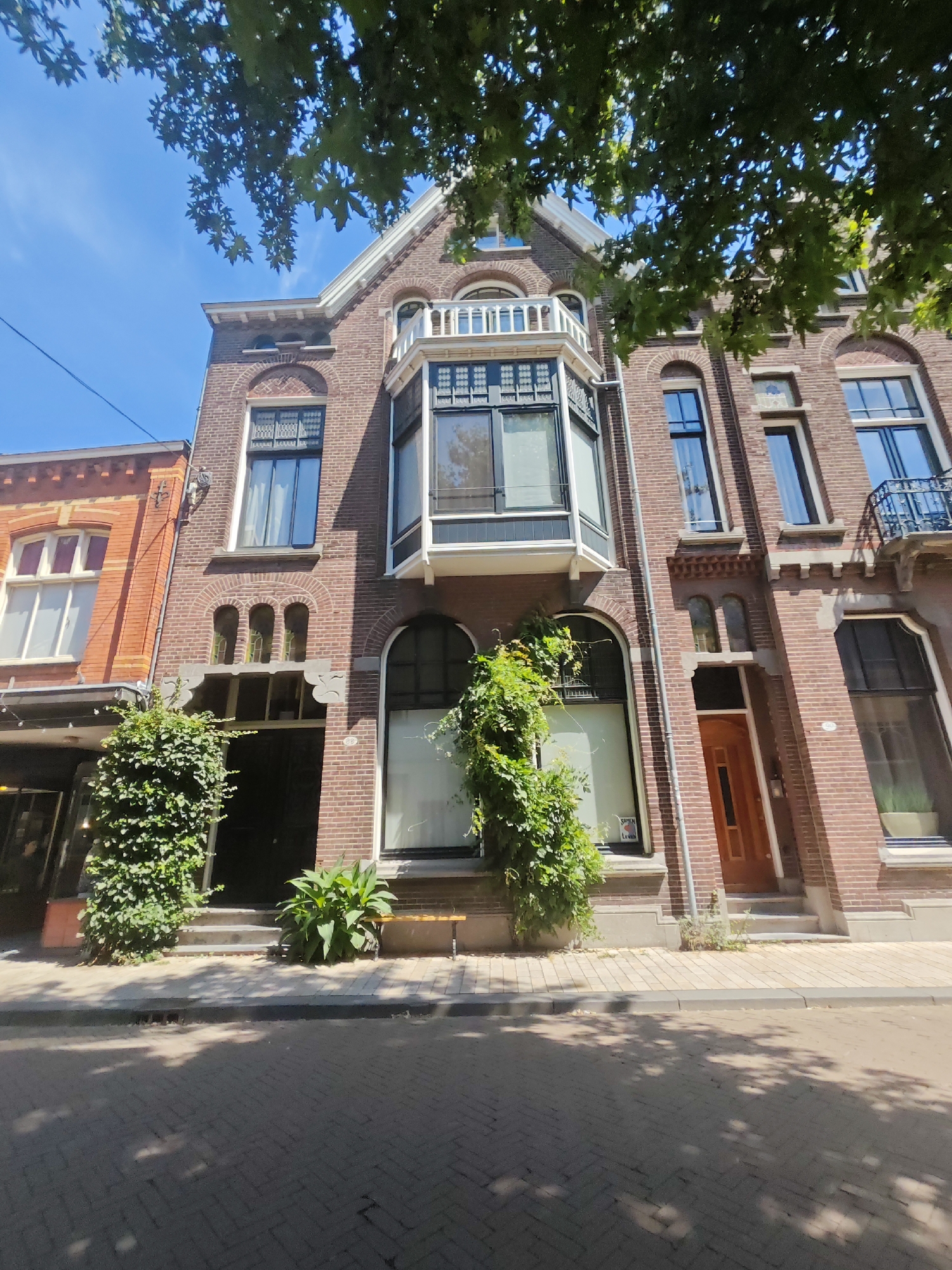 This is a low-angle, full shot image of the facade of a brick building with several windows and an entrance. The building is located in the city of Tilburg, Netherlands. The building is brown brick, with decorative white trim around the windows and roofline. The facade has a bay window on the second story and a balcony on the third story. There are several green plants growing on the exterior of the building, including ivy that climbs up the front of the building and a large potted plant in front of the entrance. The sidewalk in front of the building is paved with brick, and the street is also paved with brick. There are trees overhead, casting shadows on the sidewalk and street. The sky is blue and clear. No people are visible in the image.