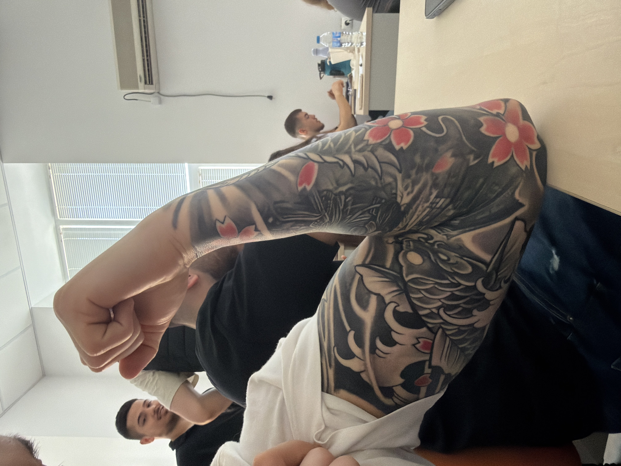 A close-up, rotated, slightly high-angle shot depicts a forearm prominently featuring a tattoo of a koi fish and cherry blossoms. The person wearing the tattoo is seated at a light-colored table in what appears to be an office or conference room. Other people are visible in the background, seated at the same table. An air conditioning unit hangs from the ceiling. The image's location in Plovdiv, Bulgaria, cannot be determined from the visual information provided.