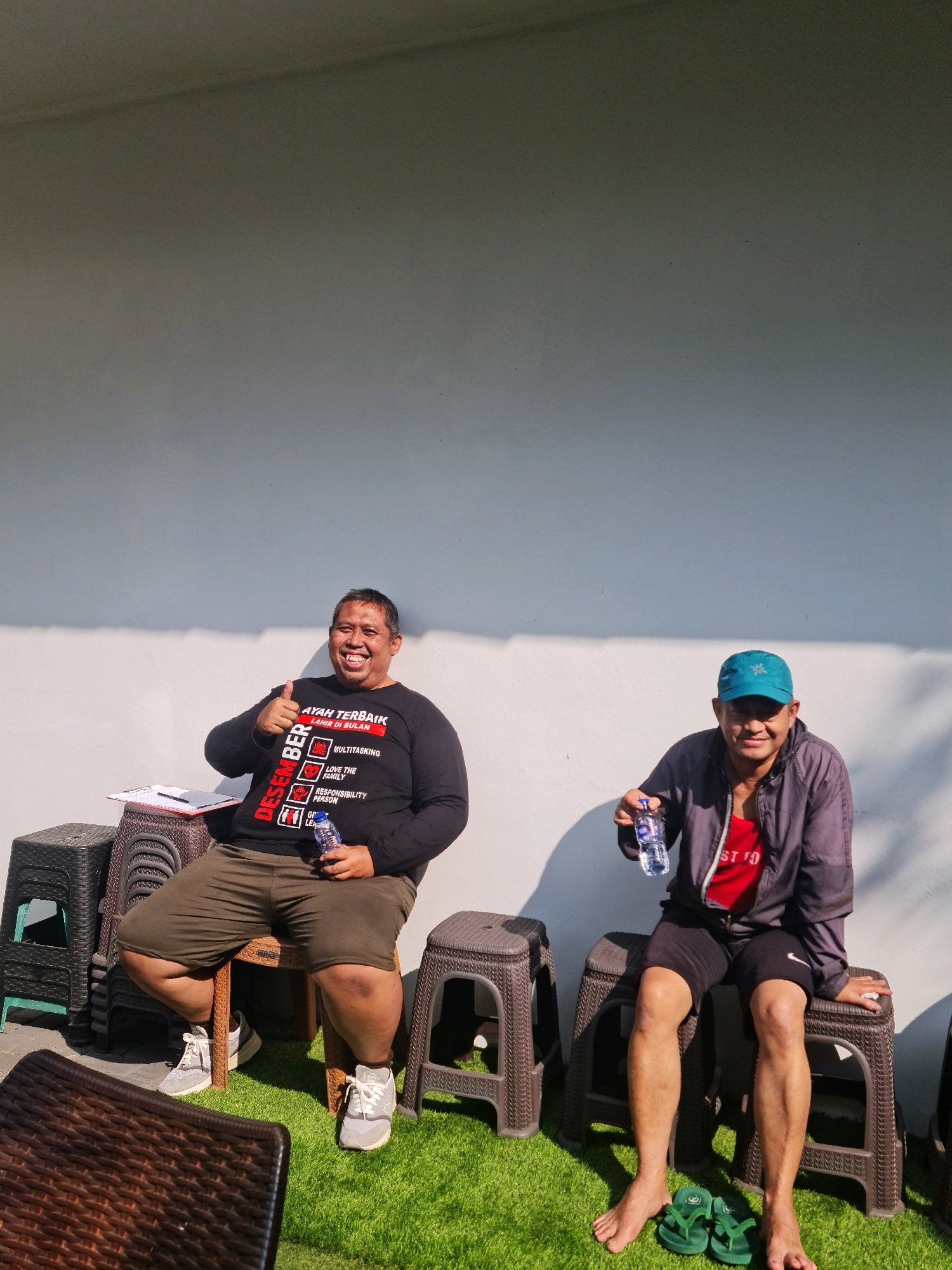 Two adult men are seated on plastic stools outdoors, positioned in front of a white wall bathed in bright sunlight, casting long shadows. The ground is covered in artificial turf.
The man on the left, with darker skin and a smiling expression, is giving a thumbs-up gesture with his left hand. He is wearing a black long-sleeved shirt with red text that reads "AYAH TERBAIK LAHIR DI BULAN DESEMBER" (Best Father Born in December) and lists qualities like "MULTITASKING," "LOVE THE FAMILY," and "RESPONSIBILITY PERSON." He is also wearing olive green shorts and light-colored athletic shoes. A bottle of water is in his right hand. A clipboard with a pen rests on a stack of plastic stools next to him.
The man on the right, with lighter skin and wearing a turquoise baseball cap, is holding a bottle of water in his left hand. He has a subtle smile. He is dressed in a gray jacket over a red t-shirt, black shorts, and is barefoot, with a pair of green flip-flops placed on the artificial turf in front of him.
The scene suggests a casual gathering or break, possibly in a park, outdoor cafe, or residential backyard in Semarang, Indonesia, given the presence of artificial turf and the men's attire. The harsh sunlight indicates it is daytime, likely midday or afternoon.
