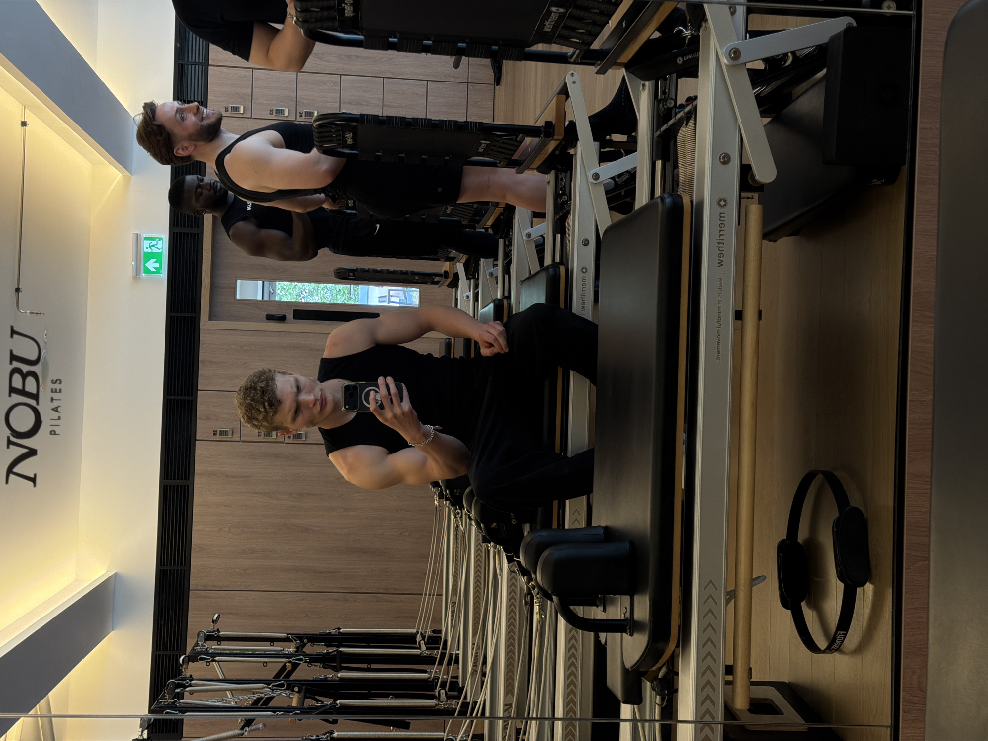 The image depicts the interior of a Nobu Pilates studio, likely in London, United Kingdom. The studio features pilates reformer machines aligned in a row. Several individuals are present, including a man taking a selfie on one of the reformers. He is wearing black athletic clothing. Another man is positioned on a reformer behind him. Behind him there is a locker with multiple doors. The background features wood-paneled walls and mirrored surfaces. The studio has minimalist branding and lighting. Headphones are hanging on the wall.