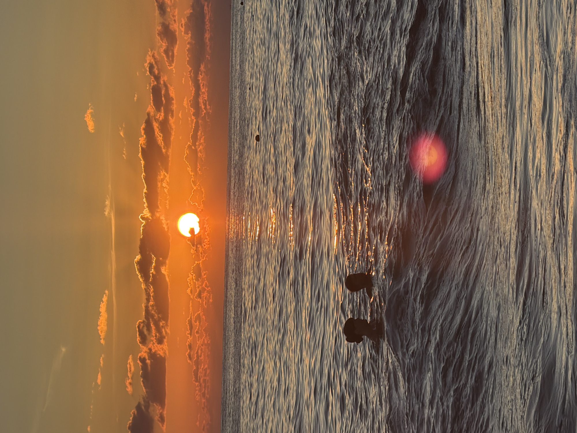 The image depicts a serene seascape at sunset, likely along the coast of Sochi, Russia, given the warm, orange-hued sky characteristic of sunsets in the region. Two individuals are partially submerged in the gently rippling water, their heads and shoulders visible above the surface. Their presence suggests a recreational activity such as swimming or wading. The sun, a brilliant orb, is setting on the horizon, casting a golden reflection across the water. Scattered clouds, illuminated by the setting sun, add texture and depth to the sky. In the distance, a few buoys are visible, indicating the presence of a designated swimming or boating area. The overall atmosphere is peaceful and tranquil, with the natural beauty of the sunset dominating the scene.