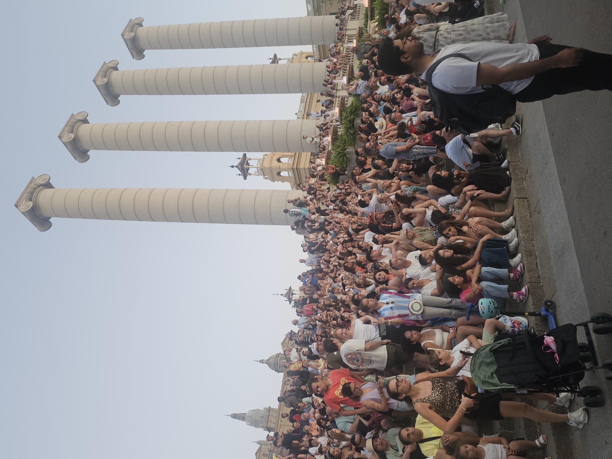 The image captures a large crowd of people gathered on broad, tiered stone steps in front of four imposing classical columns. The columns, with their distinctive spiral capitals, suggest a significant architectural landmark. In the background, several buildings with domes and ornate facades are visible, including a prominent structure with a spire, all bathed in the soft light of what appears to be late afternoon or early evening. The people are dressed in casual summer clothing, with many sitting on the steps, watching something off-camera. Some are standing, and a few young children are present, one in a stroller. The overall atmosphere is one of a public gathering, possibly for a performance or event. This scene is identifiable as the Palau Nacional, located on Montjuïc hill in Barcelona, Spain, with the four columns belonging to the Four Catalan Capitals monument.