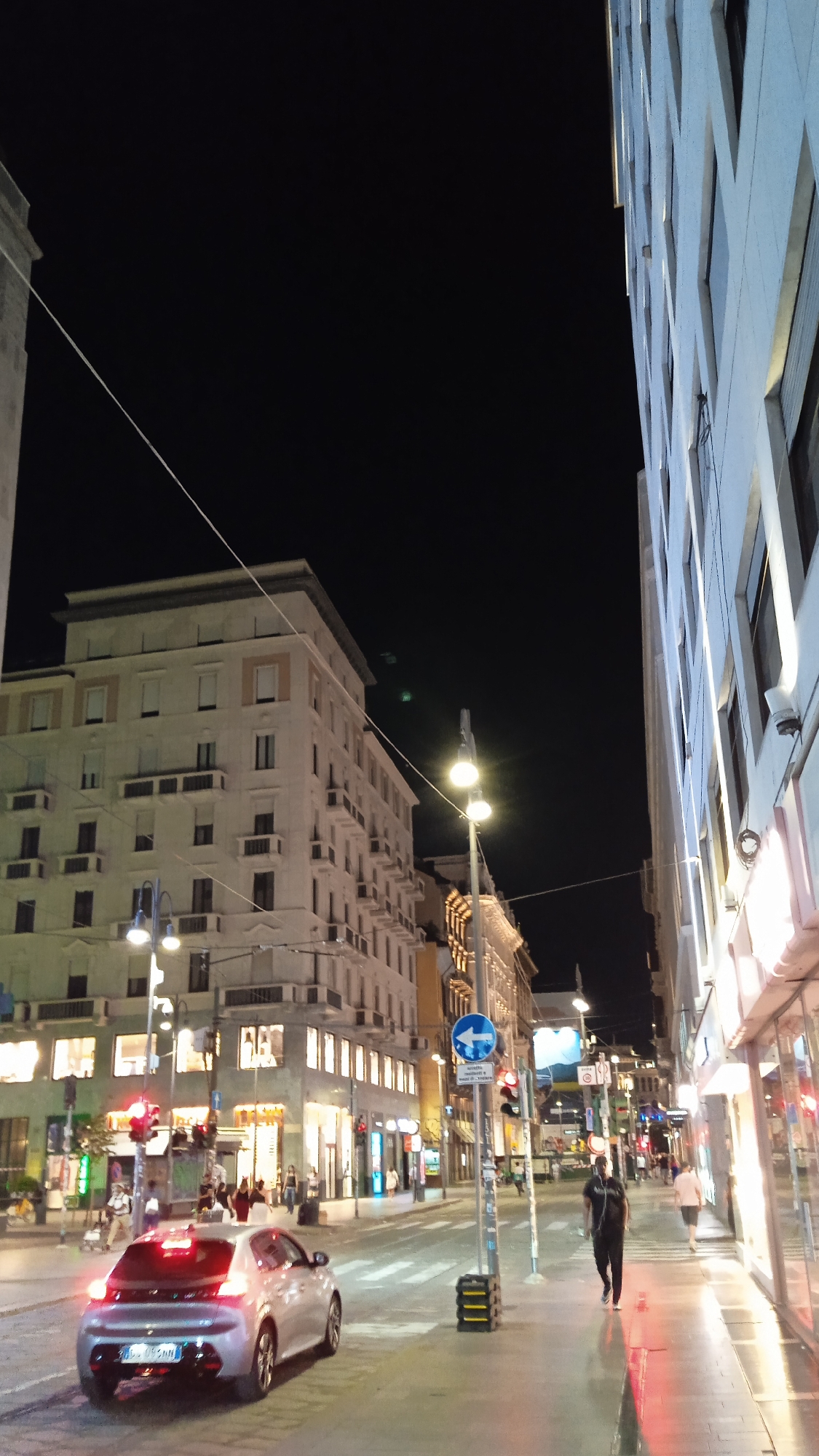The image depicts a street scene in Milan, Italy, at night. A silver Peugeot hatchback is stopped at a traffic light on the left side of the frame. In the background, a multi-story building with illuminated windows lines the street, and a modern building with a patterned facade is visible on the right. Pedestrians are walking along the sidewalk, and streetlights illuminate the scene. A traffic sign with a blue arrow indicating a left turn is visible. The sky is dark, suggesting it is late evening or night.