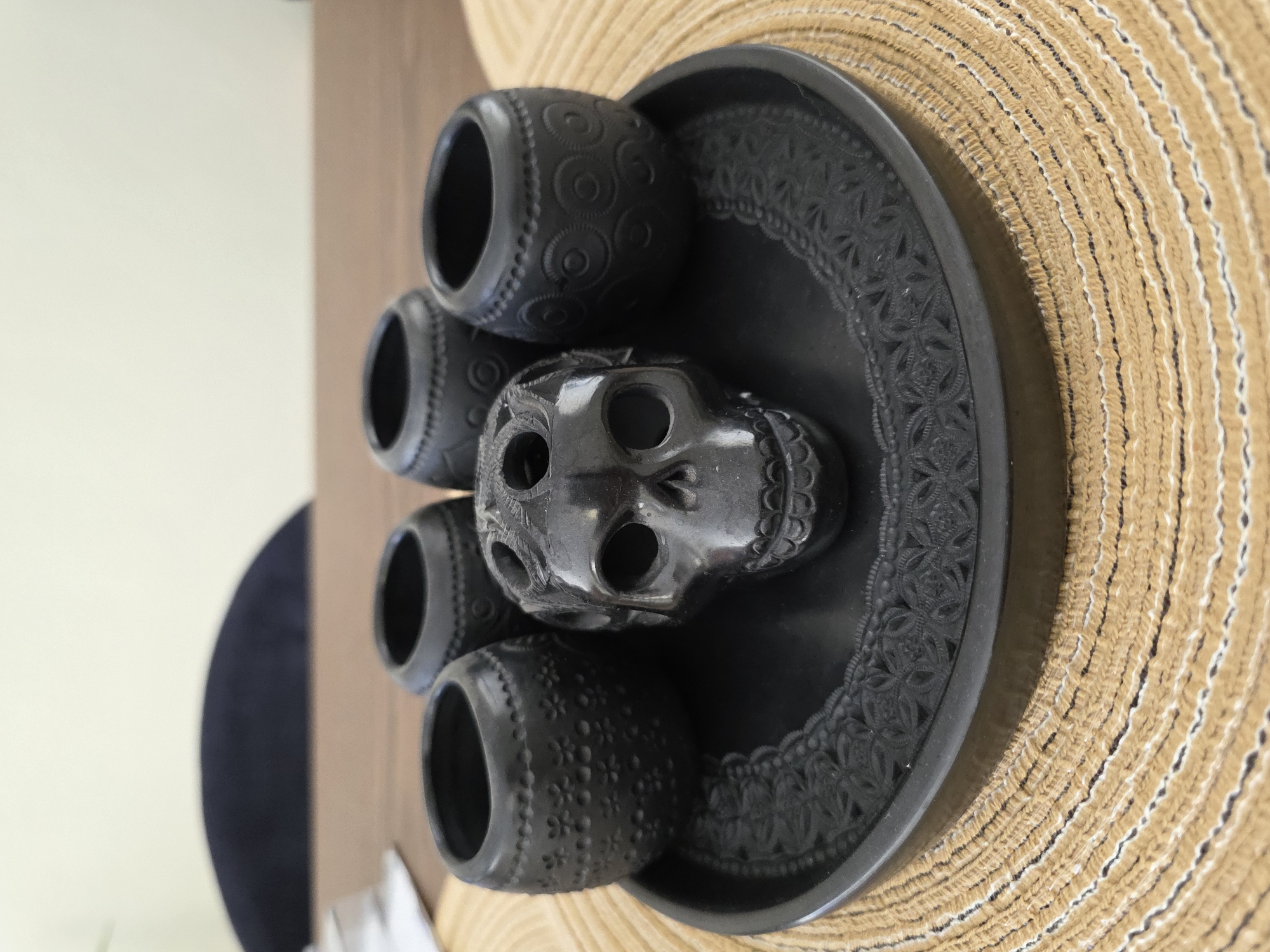 The image displays a decorative arrangement of black polished ceramic objects, set indoors on a textured, woven mat. The main subjects are a stylized ceramic skull, four small cylindrical cups or vessels, and a round tray, all crafted from the same dark material.
The skull, centrally placed on the tray, features intricately carved details, including prominent eye sockets and nasal cavities. Its surface exhibits a subtle sheen, reflecting light. Surrounding the skull are four identical-sized vessels, each adorned with distinct carved patterns; one vessel shows a repetitive spiral motif, while another displays a floral design. The round tray supporting these pieces is also highly decorated, with an ornate, repeating pattern carved into its rim.
These black ceramic items rest on a light brown, woven mat, characterized by its concentric circular pattern and visible texture. The background consists of a light, possibly cream-colored wall, with a blurred, dark blue or black shape partially visible on the left, suggesting other furniture or decor. The scene is brightly lit, emphasizing the intricate details and smooth finish of the pottery. There is no activity occurring, and no visible text. The style of the pottery is characteristic of traditional Mexican craftsmanship, in the context of Puebla, Mexico.