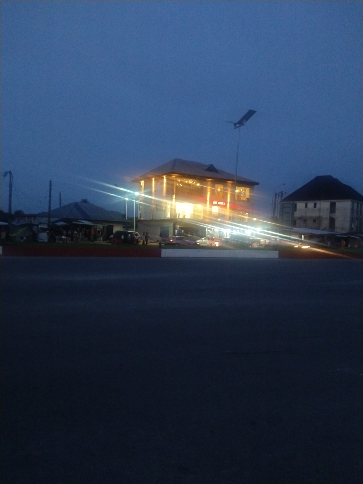 The image shows a building with the name "THE NEST" illuminated at night in Uyo, Nigeria. The building appears to be a commercial establishment. The sky is a deep blue, suggesting it is either dusk or dawn. A streetlight is visible to the right of the building. Several cars are parked or driving in front of the building, creating light trails. People are gathered around what appears to be a smaller structure or stall on the left of the main building. The foreground is dominated by a dark road or pavement. A two-story building with a dark roof is on the right side.