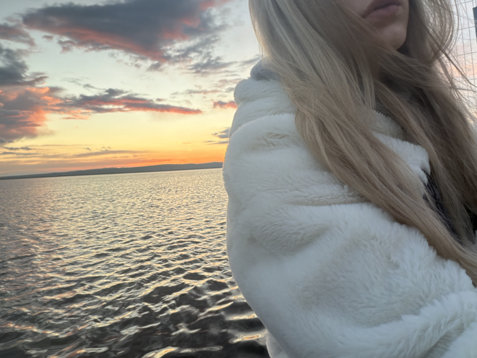 The image shows a woman standing near a body of water, possibly a lake or a sea, during sunset. The sky is filled with clouds tinged with orange and pink hues. The water surface is rippled, reflecting the colors of the sky. The woman is wearing a white, fluffy jacket. Her long, blonde hair falls over her shoulder. A fence is barely visible in the background near the woman's head. The exact location in the city of Unknown, Unknown, cannot be determined from the image.