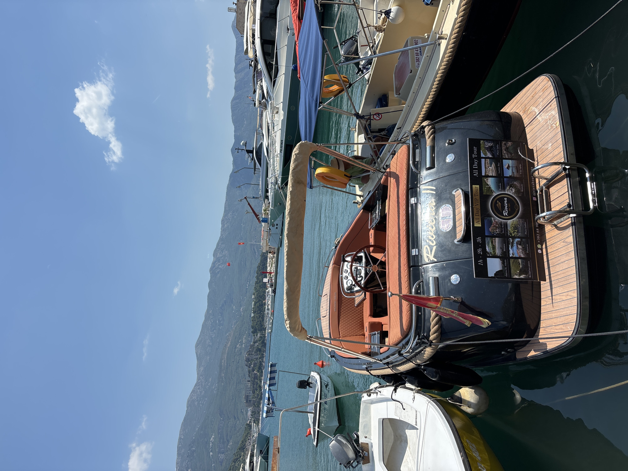 A black motorboat with orange upholstery is docked in a harbor in Budva, Montenegro. The boat's stern displays a sign advertising "All Day VIP Tour" and "All Day Tour," with images of local attractions and contact information. A small red flag is attached to the stern. The boat is tethered to a larger yellow boat on its right. In the background, other boats and yachts are moored, and mountains rise behind the city. The sky is clear blue with a few scattered clouds.