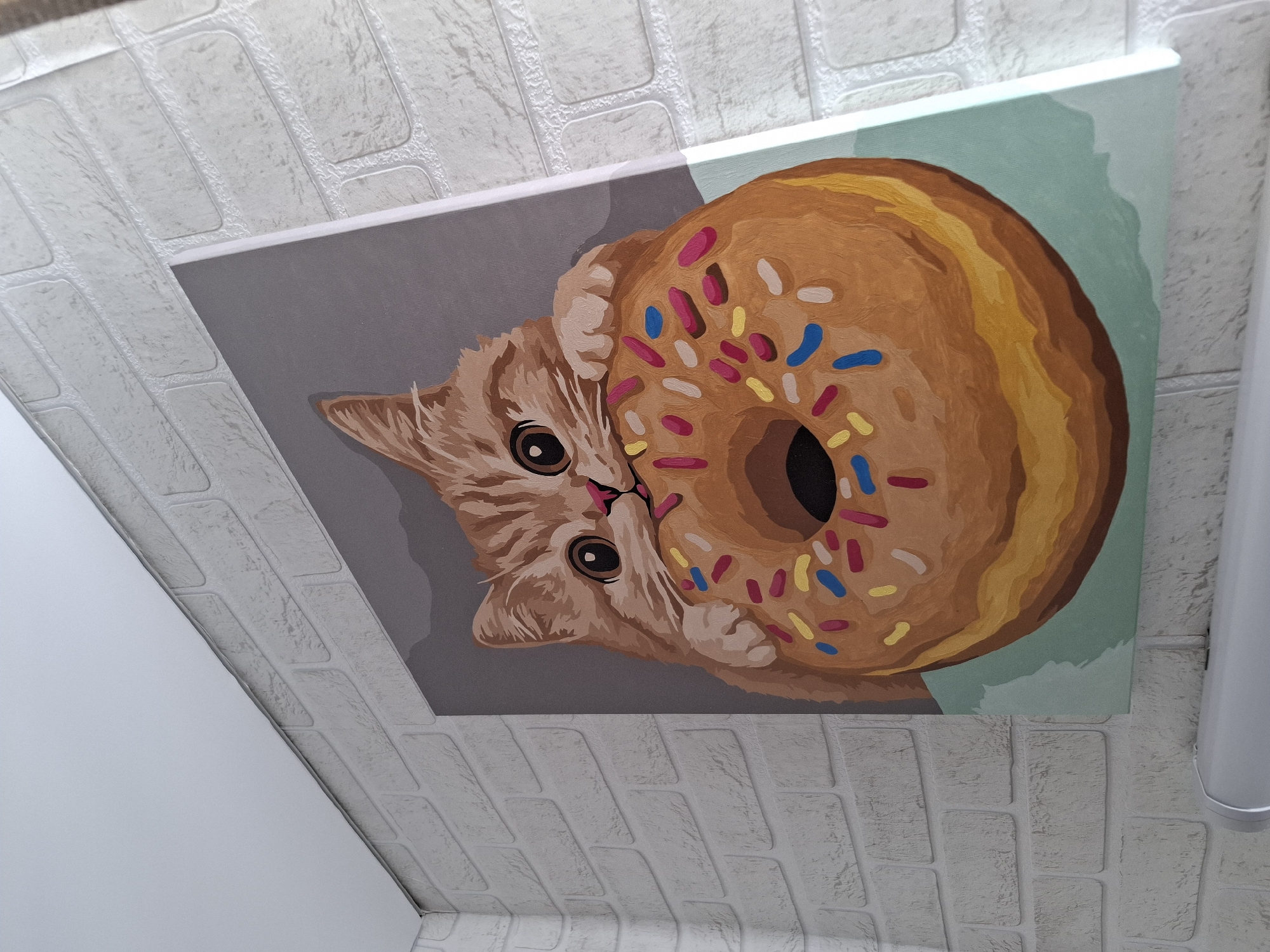 A canvas painting featuring a stylized ginger or light brown tabby cat and a large, frosted donut is prominently displayed. The cat has large, dark eyes and a pink nose, with its paws seemingly resting on the donut. The donut is a warm tan-brown with colorful sprinkles in red, blue, yellow, and white, and a distinct central hole. The painting employs a graphic, somewhat flat art style, with the cat set against a grey background and the donut against a light teal/mint green background.
The painting is mounted on an interior surface, likely a wall or ceiling, which is covered with light grey or white textured panels that resemble embossed brickwork or stone blocks. A white, cylindrical light fixture is partially visible on the right side of the frame, also attached to this textured surface. The image is taken at an upward angle, showing the painting fixed to the overhead or upper wall space.
No specific activity or event is occurring beyond the static display of the artwork. The scene is brightly lit, likely by indoor artificial light. The painting evokes a whimsical and cheerful mood due to its subject matter and colorful palette. There is no visible text in the image. This media file was captured in Амзя, Russia.