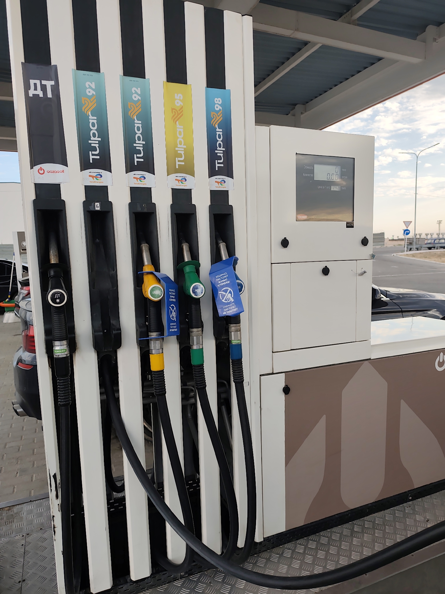 The image shows a close-up view of a multi-pump gasoline dispenser at a gas station in Ömirzaq, Kazakhstan. The dispenser has five nozzles, each labeled with a different fuel type: "ДТ" (diesel), "Tulpar 92" (twice), "Tulpar 95", and "Tulpar 98". The "Tulpar" brand appears to be associated with TotalEnergies. The dispenser features a digital display showing "0.00" for cost and quantity, and the price per liter as "242". The nozzles are black, yellow, green, and blue, with some having attached blue warning labels. In the background, a dark-colored car is partially visible, along with a road, streetlights, and a cloudy sky. The ground is covered with a metal checkered plate. The overall scene is a typical view of a modern gas station.