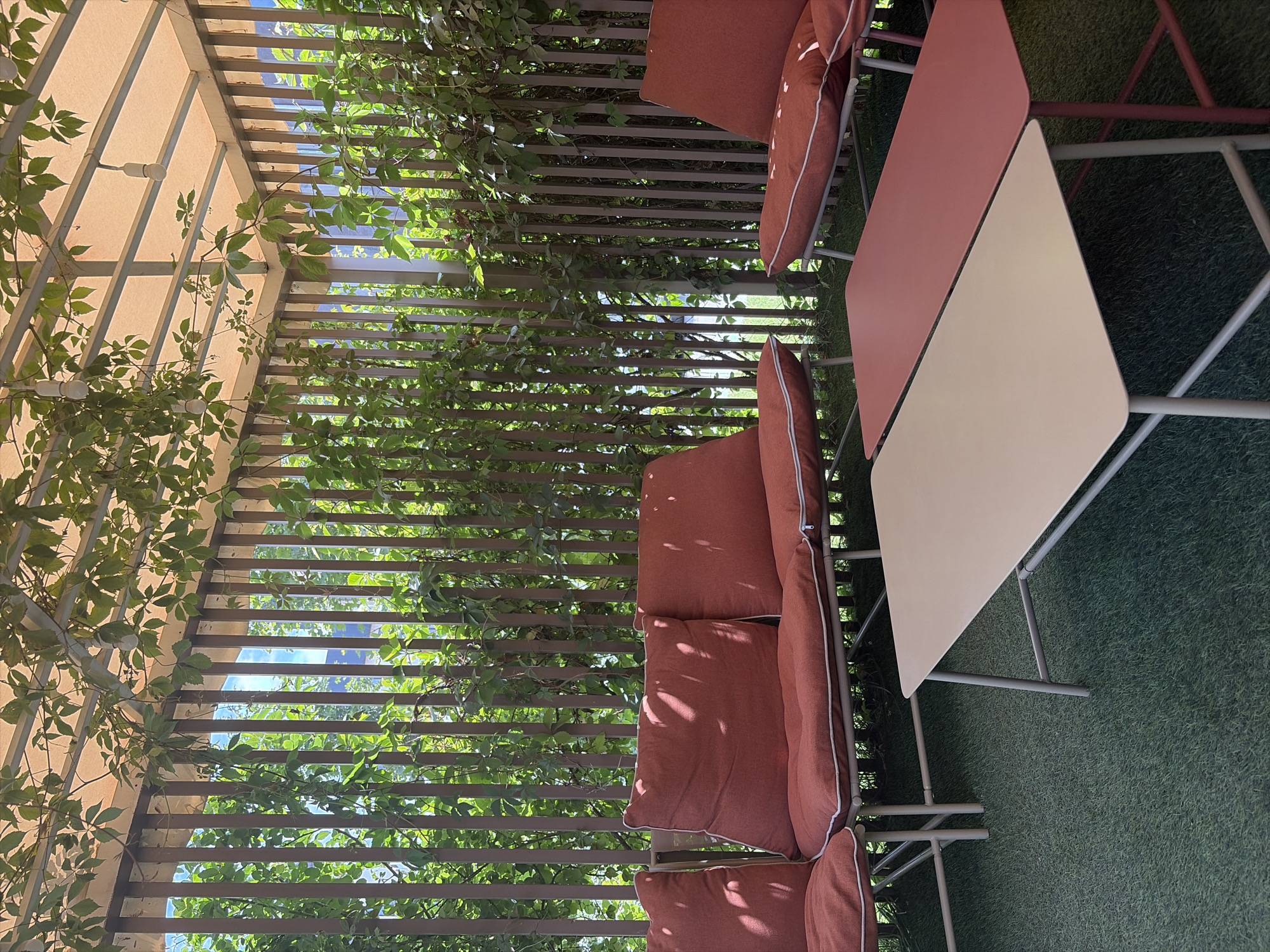 The image depicts an outdoor seating area, likely a patio or balcony, covered by a light-colored canopy with hanging lights. A wooden slatted fence provides a backdrop, intertwined with lush green vines. In the foreground, there are two contemporary coffee tables, one a light beige and the other a dusty rose, placed on a green artificial turf floor. Behind the tables are rust-colored upholstered outdoor sofas and chairs. The scene appears to be a serene and comfortable spot for relaxation. The presence of vines and the outdoor setting suggests a location in Bishkek City, Kyrgyzstan, possibly a cafe, restaurant, or residential balcony.