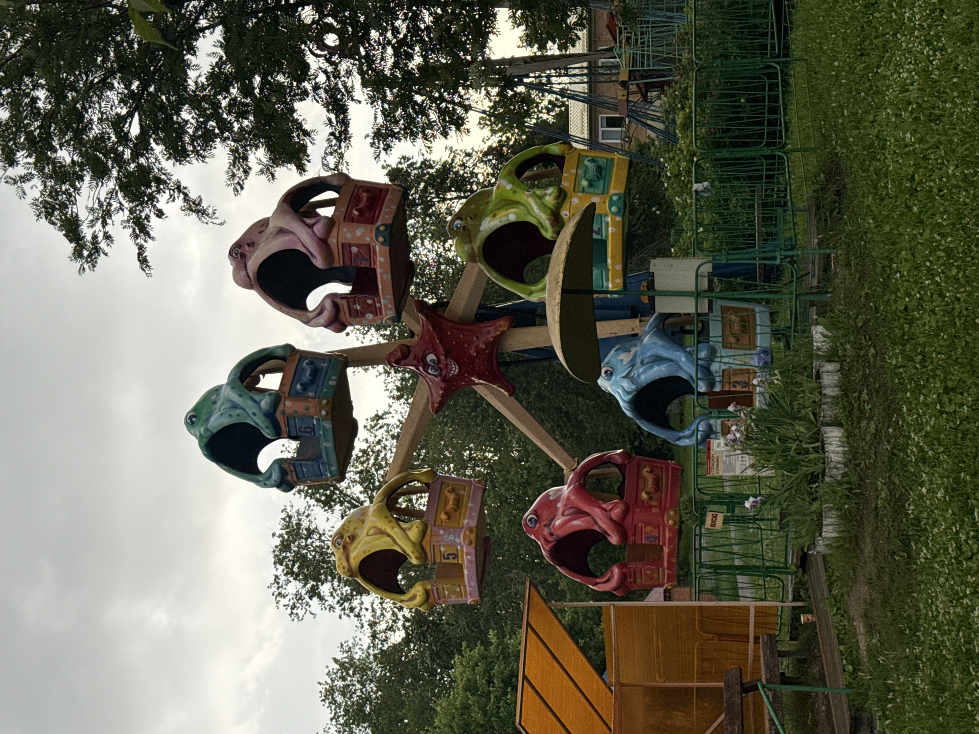 The image shows a small, faded amusement park ride, possibly a carousel or a miniature Ferris wheel, with several gondolas designed in the shape of octopus heads. The ride is situated in an outdoor, grassy area, with trees in the background and a cloudy sky overhead. To the left, there is an orange awning and a wooden bench. Fencing encloses the ride. In the background, a building and other amusement park structures are visible. The overall impression is one of a slightly neglected, old-fashioned amusement park. The ride appears to be unoccupied. The image does not contain any specific details to confirm its location in Chusovoy, Russia, as it is a generic scene of an amusement park.