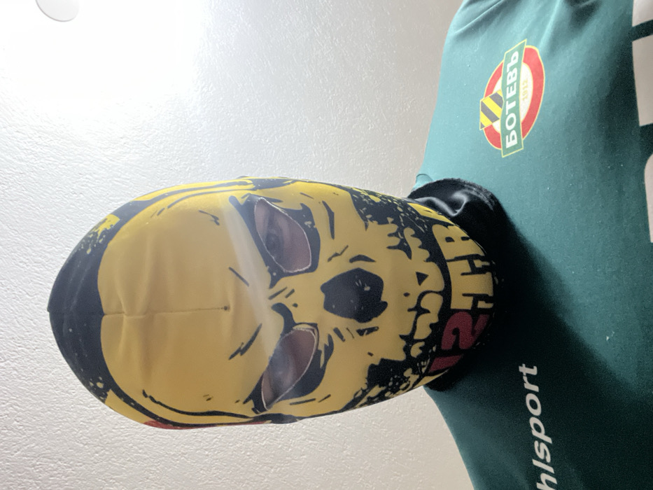 A close-up, chest-up image features an individual wearing a yellow and black balaclava with a skull motif, revealing only the eyes through cutouts. The individual wears a dark green athletic shirt that displays "uhlsport" in white text on the viewer's left and a circular logo on the viewer's right. The logo is composed of a red outer ring, a green inner field, yellow and black diagonal stripes at the top, and the white Cyrillic text "БОТЕВЪ 1912". The individual faces forward against a plain, light-colored interior wall, illuminated by a light source from the upper right. This scene is located in Sedlartsi, Bulgaria.
