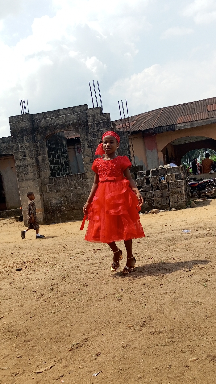The image presents an outdoor scene in what appears to be a rural or semi-urban setting, identified as Agburu Uke, Nigeria. The ground is unpaved, covered in dry, light brown earth with visible footprints and sparse vegetation.
The primary subject is a young girl, estimated to be between 5 and 8 years old, positioned centrally in the foreground. She is adorned in a bright, layered red party dress with short sleeves and intricate floral or beaded details on the bodice. A matching red bow cinches her waist, and she wears a red headscarf or headband. Her feet are shod in golden or metallic heeled sandals with decorative bows. The girl appears to be mid-stride or in a slight hop, looking towards the viewer with a neutral to slightly smiling expression.
In the mid-ground to the left, a younger boy, possibly a toddler, walks away from the camera, wearing a striped shirt and shorts. The background features several buildings constructed from concrete blocks. On the left, a structure appears incomplete, with rebar rods extending upwards. To the right, another building has a corrugated metal roof and an archway. Through this archway, indistinct figures are visible, some seated, and a parked motorcycle can be seen.
The time of day is bright daylight, indicated by the well-lit scene and shadows, with a sky full of white and grey clouds. The overall atmosphere suggests a special occasion for the girl, contrasting with the rustic and unpaved environment. There is no visible text in the image.