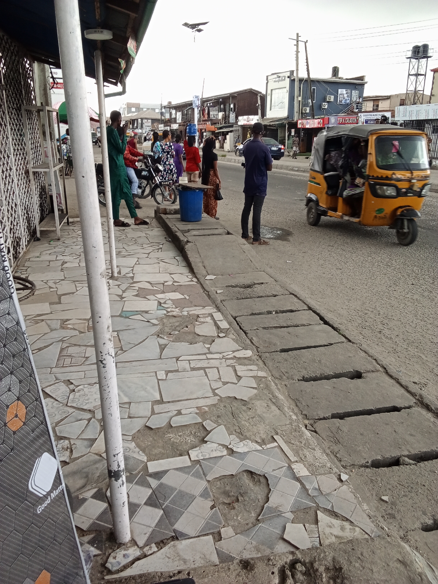 The image shows a street scene in Ado, Nigeria. In the foreground, a dilapidated sidewalk with broken tiles runs alongside a concrete drainage ditch with missing or broken sections. A sign partially visible in the bottom left corner advertises "Good Mattr." People are standing on the sidewalk and along the roadside. Some individuals appear to be waiting near a blue plastic barrel. In the background, there is a small yellow three-wheeled vehicle, a "keke", on the road. The street is lined with buildings, some of which have signage for businesses. Power lines and a communications tower are visible in the distance. The overall atmosphere suggests a busy, but somewhat rundown urban environment.