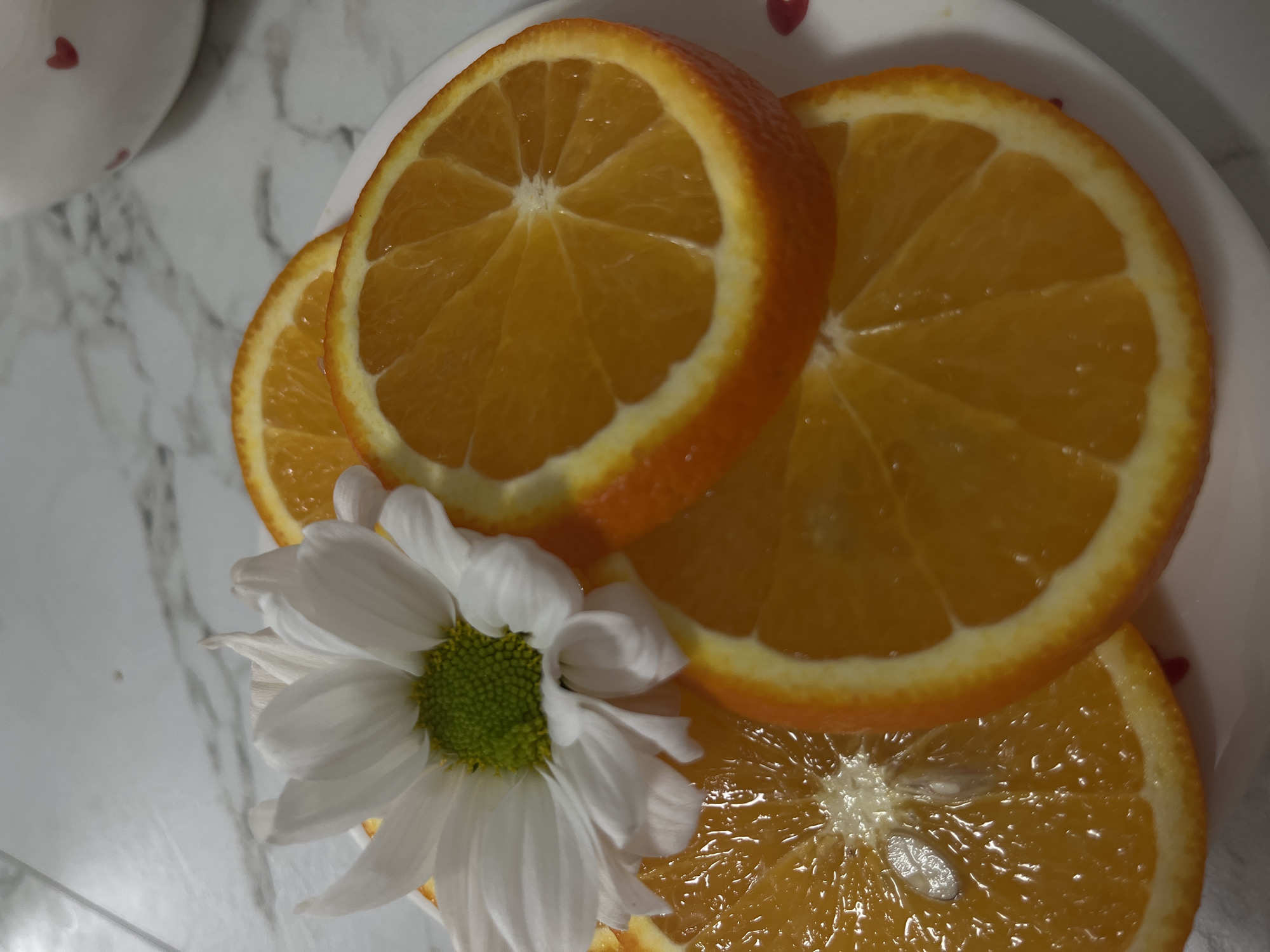 A close-up shot presents a plate of sliced oranges, adorned with a white daisy. The oranges, cut into circular slices, display a vibrant orange hue with the segmented flesh clearly visible. The daisy, positioned near the bottom left of the image, features white petals surrounding a green center. The plate rests on a surface that appears to be a white marble countertop or table. The background is blurry, but a white object with red hearts is visible in the top left corner. The location of Chaikovsky, Russia, is not evidenced in the image.