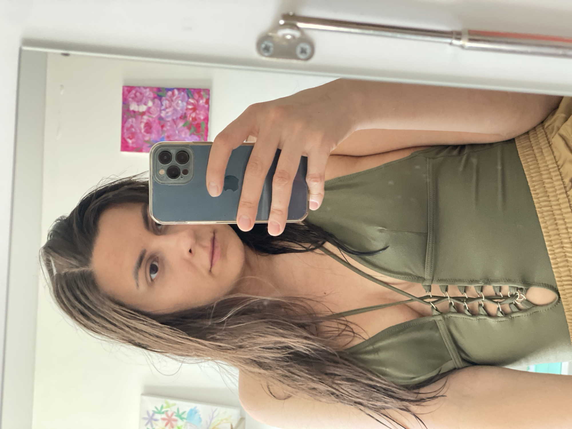A selfie taken in a bathroom mirror shows a woman with long, damp brown hair and fair skin, holding a blue smartphone in her right hand. The phone partially obscures her face. She is wearing an olive green top with decorative metal rings and eyelets at the bust and tan, gathered-waist shorts. The mirror reflects details of the bathroom, including a silver towel bar at the top, a pink floral painting on the wall, and a partially visible floral artwork at the bottom of the frame. The location is Unknown, Unknown.