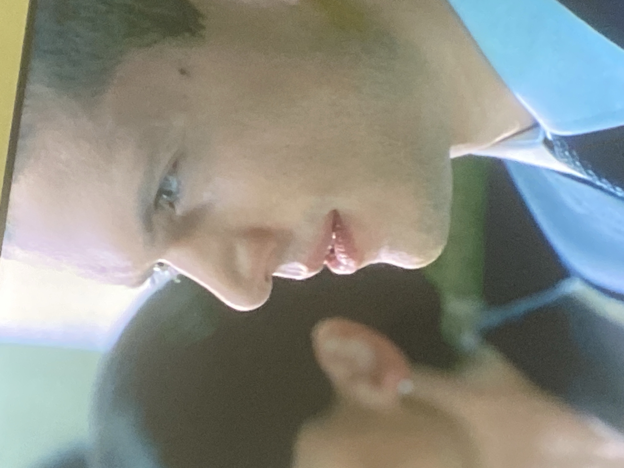 A close-up, rotated image captures a section of a person's face, specifically from the eye down to the chin, and part of their neck and shoulder, alongside a blurred hand in the foreground. The person, who appears to have light to medium skin tone, is seen in profile with their head tilted. A light-colored eye (possibly blue or grey) is visible, and there's a small dark spot on the cheek. Short, dark hair is visible at the top left. The lips are slightly parted, revealing some teeth. Below the chin, the outline of a jawline with what appears to be light stubble is discernible.
On the right side of the frame, part of a light blue collar and a dark, possibly patterned, tie or strap are visible, suggesting formal or semi-formal attire. In the lower right corner, a blurry hand is present, with a ring visible on one of the fingers, possibly the pinky or ring finger. The background is indistinct and out of focus, showing blurry patches of green and dark tones. The lighting is soft, and there are no clear indications of the time of day or specific weather conditions. No visible text is present. The overall scene gives the impression of a moment captured during speech or deep thought, likely from a video or television screen due to the image quality. This scene is contextualized as being in Gosa, Nigeria.