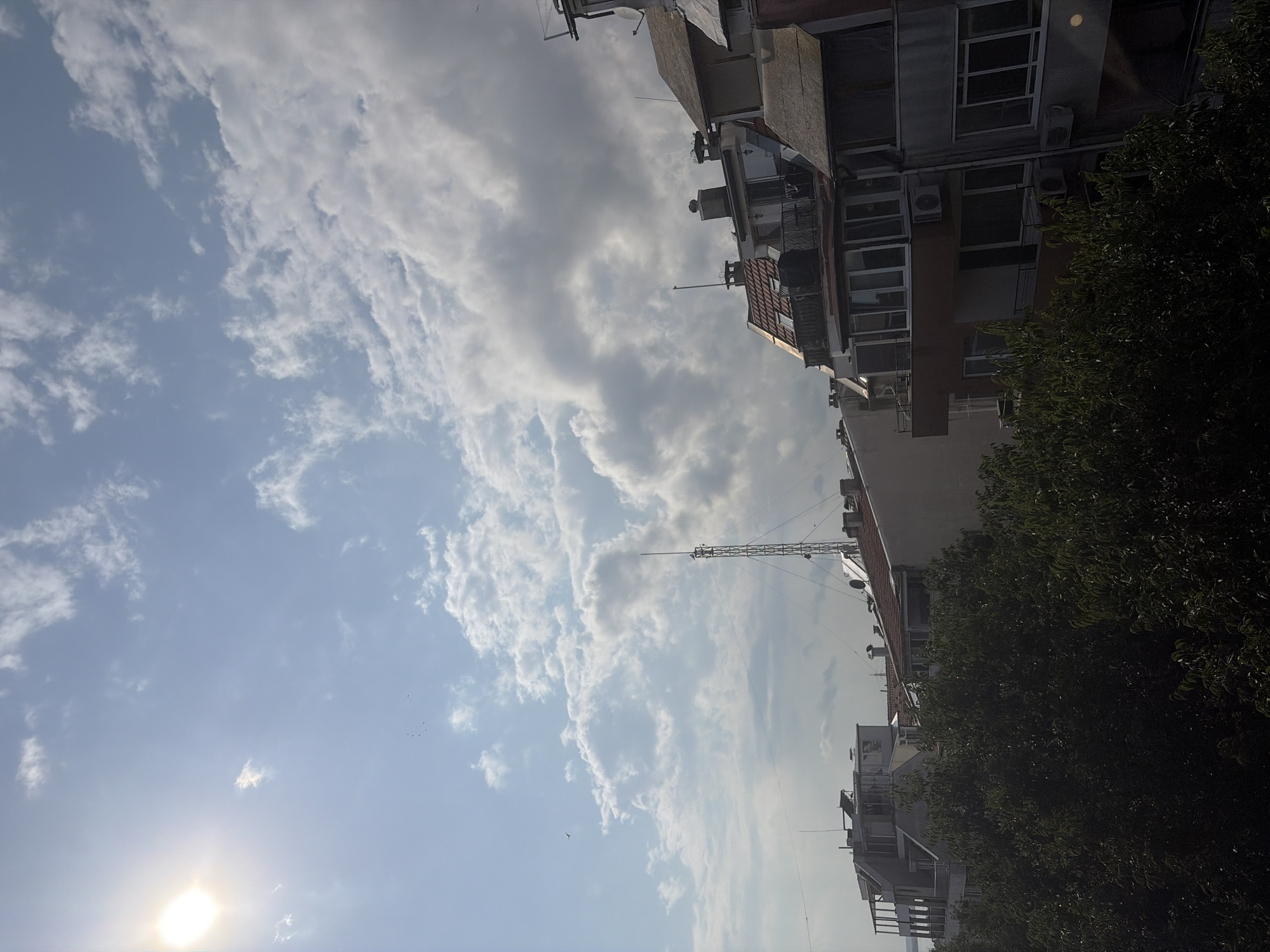 The image depicts a cloudy sky over a city skyline, likely Burgas, Bulgaria, with several buildings visible. The sun is shining brightly on the left side of the frame, partially obscured by clouds. In the foreground, dense green foliage obscures the lower portion of the scene. The middle ground features rooftops of several buildings, characterized by their varied architecture and materials, including tiled roofs and concrete structures. A prominent telecommunications tower with an antenna stands tall among the buildings. Some of the buildings have visible balconies, windows, and air conditioning units. A few birds are seen flying in the sky. The overall impression is of an urban environment under a dynamic sky.