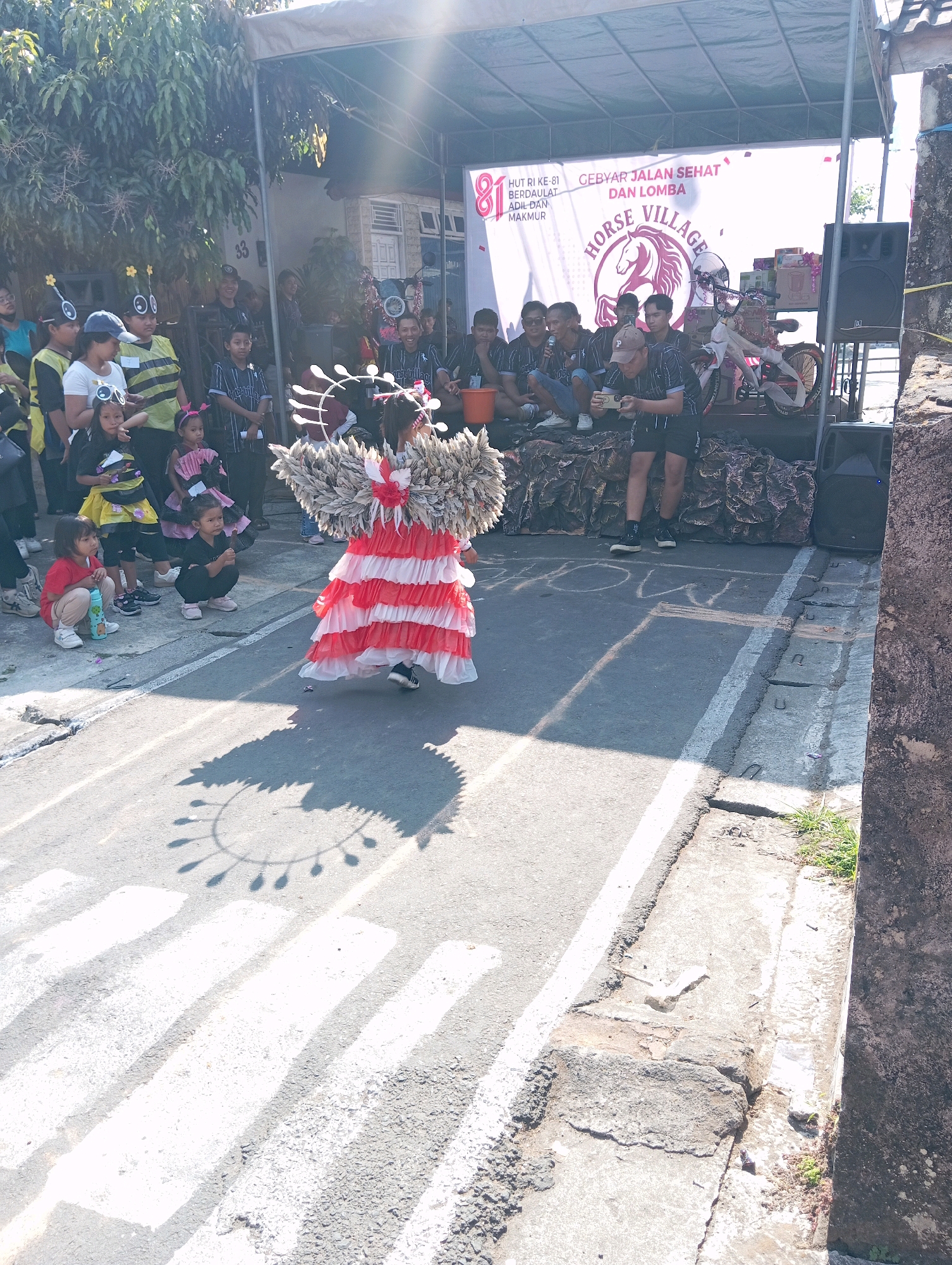 The image depicts an outdoor event in what appears to be a street in Magelang, Indonesia. A young person in a vibrant red and white ruffled costume, resembling a flower or a bird's plumage, is the central focus, walking away from the camera. This individual is adorned with a large headdress featuring white feather-like elements. Their shadow, cast on the asphalt, is an intricate circular pattern with dangling elements.
Behind the costumed individual, a crowd of people is gathered. Some are spectators, including young children squatting on the ground, while others appear to be participants or organizers. Several children are dressed in bee-themed costumes, complete with yellow and black stripes and antennae headbands.
In the background, a stage or display area is set up under a canopy. A large banner with the text "HUT RI KE-81 CEBYAR JALAN SEHAT DAN LOMBA HORSE VILLAGE" is prominently displayed, indicating a celebratory event possibly related to Indonesia's Independence Day (HUT RI) and a healthy walk/run and competition. A bicycle, adorned with gifts and ribbons, is displayed on the stage, likely a prize. Audio equipment, including speakers, is also visible.
The environment suggests a sunny day, with bright sunlight casting strong shadows. The street itself has white pedestrian crossing markings. The surrounding area includes buildings and trees, characteristic of an urban or semi-urban Indonesian setting. The overall scene captures a lively community event, possibly a parade or festival.
