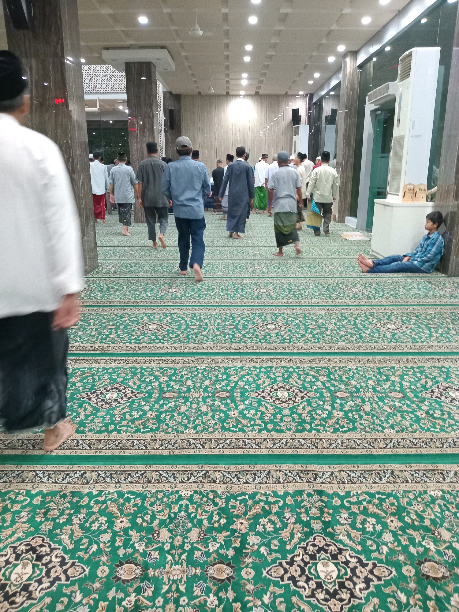 The image depicts the interior of a mosque in Randuagung, Indonesia, characterized by its vast green carpet with intricate floral patterns. Several men, dressed in traditional Islamic attire, are seen walking on the carpet. Some are barefoot, which is customary in mosques. In the background, there are large, decorative pillars and modern amenities like air conditioning units, suggesting a well-maintained and contemporary mosque. A young boy is seated on the floor near a wall, observing the activity around him. The lighting is bright, indicating artificial illumination from overhead fixtures. The overall scene conveys a sense of communal gathering and prayer within a religious space.