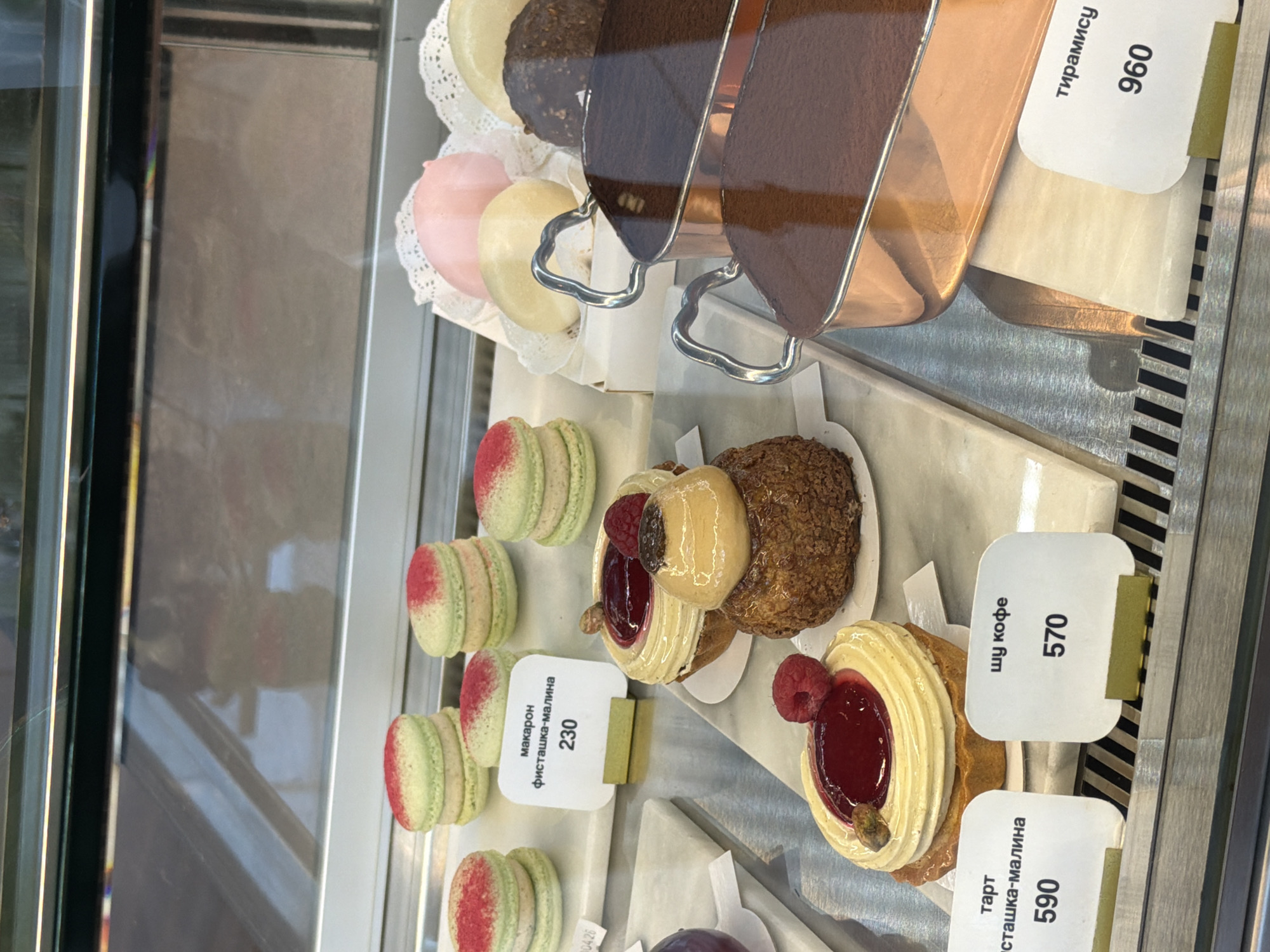 The image captures a variety of desserts displayed in a chilled glass case, likely in a pastry shop or cafe in Khimki, Russia. The scene is shot from a slightly elevated, oblique angle, with the items arranged on white and patterned surfaces. The overall impression is one of neatness and appetizing presentation.
In the foreground and mid-ground, rows of macarons with light green bodies and red specks are visible, suggesting a fruit flavor like raspberry. Next to them are individual pastries, including choux-style buns and tarts, adorned with cream, berries, and glistening glazes. One prominent tart features a yellow swirled cream, a dollop of red jam, and a fresh raspberry. Another pastry, possibly a choux, is topped with a light brown cream and a darker element. In the background, two containers hold a dark, powdered dessert, identified as Tiramisu.
Several white price tags with Cyrillic text and numbers are visible.
- A tag next to the green macarons reads "макарон фисташка-малина 230," indicating "Macaron pistachio-raspberry" priced at 230 (likely Rubles).
- A tag near the tart states "тарт фисташка-малина 590," identifying it as a "Tart pistachio-raspberry" for 590.
- Another tag reads "шу кофе 570," for a "Choux coffee" pastry priced at 570.
- The tag for the large, dark dessert in the background clearly shows "тирамису 960," for "Tiramisu" priced at 960.
A small, partial number "0426" is also visible on one of the paper liners. No people are visible, and the time of day is not discernible.
