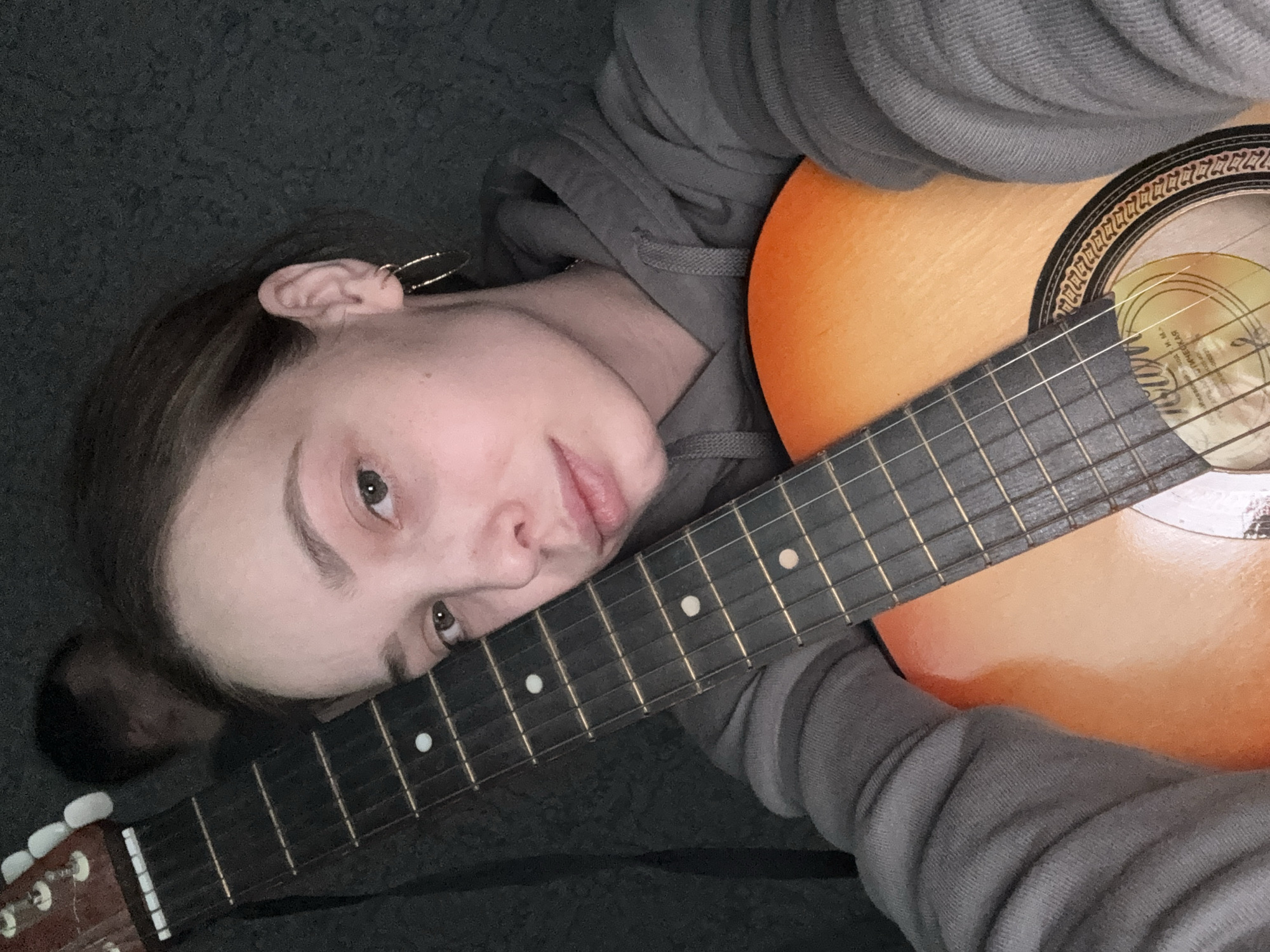 A person is lying down, with their head resting on the neck of an acoustic guitar. The guitar's soundhole is visible, as is part of its body. The person is wearing a gray hooded sweatshirt. The background is a dark, textured surface. The city of Прикамский, Russia cannot be determined from the image.