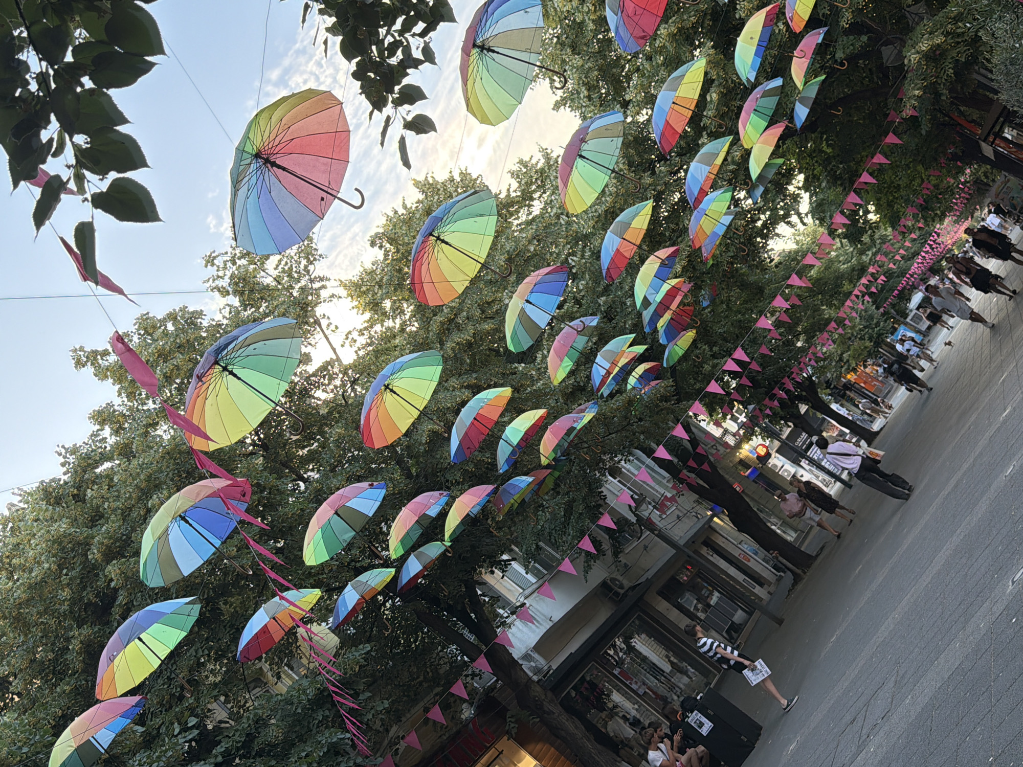This media file captures a lively outdoor scene, likely a street or pedestrian area in Burgas, Bulgaria. A prominent feature is a colorful display of numerous rainbow-colored umbrellas suspended overhead, creating a vibrant canopy. The umbrellas are arranged in a pattern, with some open and others partially closed, adding a festive and whimsical atmosphere.
Beneath the umbrellas, the scene shows a bustling pedestrian walkway lined with buildings that appear to be shops or businesses. People are visible walking along the path, suggesting a public gathering or a day of commerce. The perspective is angled, looking up towards the umbrellas and the sky, which is partially visible with some clouds. The trees with green foliage frame the scene from above, adding to the outdoor setting.
The time of day appears to be late afternoon or early evening, indicated by the warm, soft lighting and the shadows cast. The weather seems pleasant, suitable for outdoor activities. The presence of people strolling and shops suggests an active urban environment.
Notable details include small pink triangular flags strung along one side of the walkway, further enhancing the celebratory feel. Some individuals in the foreground and mid-ground are engaged in various activities, such as walking, conversing, or possibly browsing. One person in the foreground is holding up a piece of paper with some text, though it's not clearly legible. The overall impression is one of a cheerful and engaging public space.