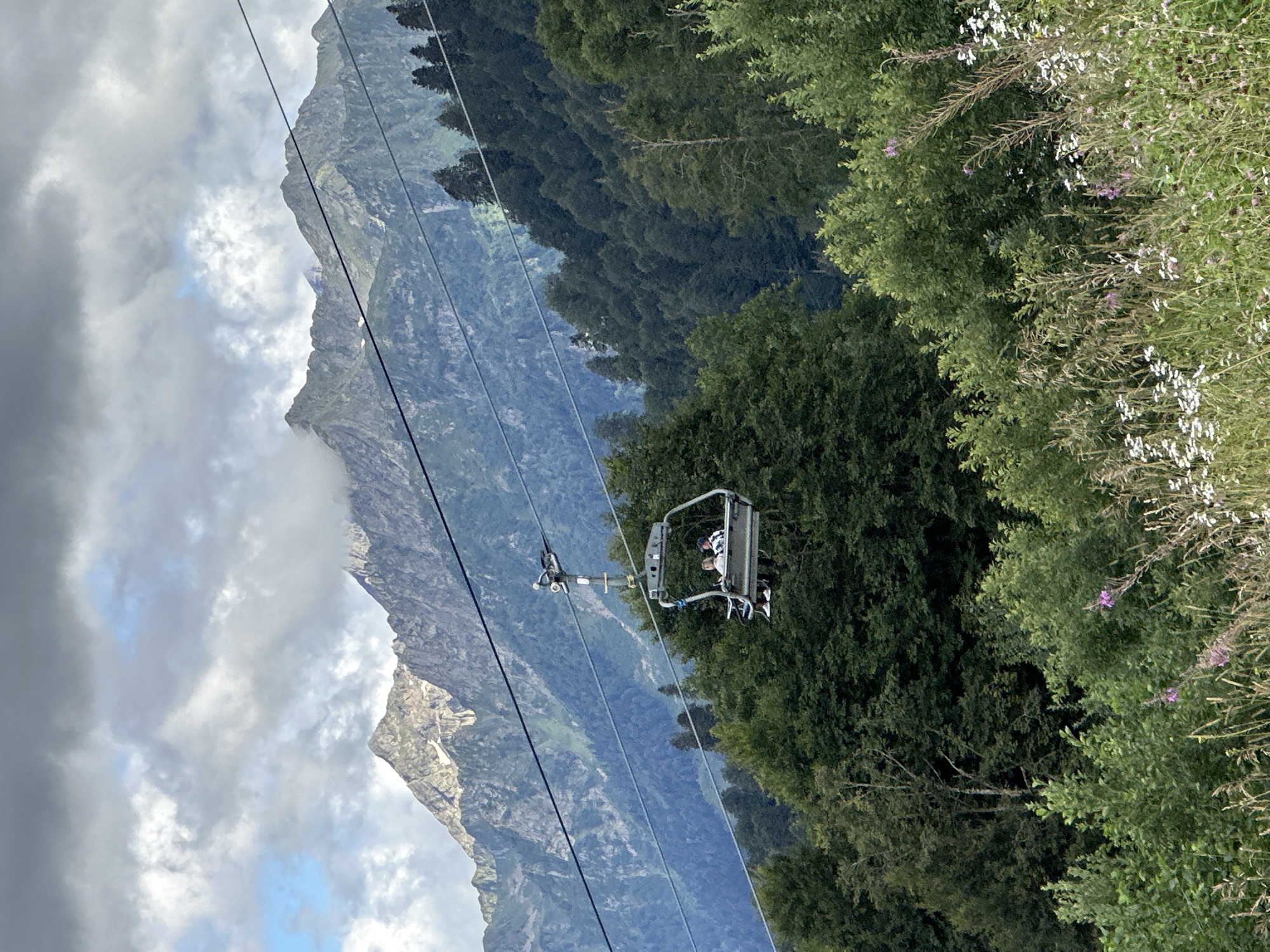 The image captures a scenic view from the ski resort town of Estosadok, Russia, likely during the summer or shoulder season, given the lush green foliage. In the foreground, wild grasses and wildflowers are abundant, with some pink and white blooms visible. Behind this, a dense forest of green trees covers the lower slopes of the mountains.
Dominating the mid-ground is a ski lift chair suspended from cables that stretch across the frame. The chair carries two individuals, one wearing a blue and white checkered shirt and the other in a lighter colored top. They are seated and appear to be enjoying the ride.
In the background, majestic mountains rise, their peaks partially obscured by dramatic clouds. Some snow patches are visible on the higher elevations, indicating a high altitude. The overall atmosphere is one of natural beauty and outdoor recreation.