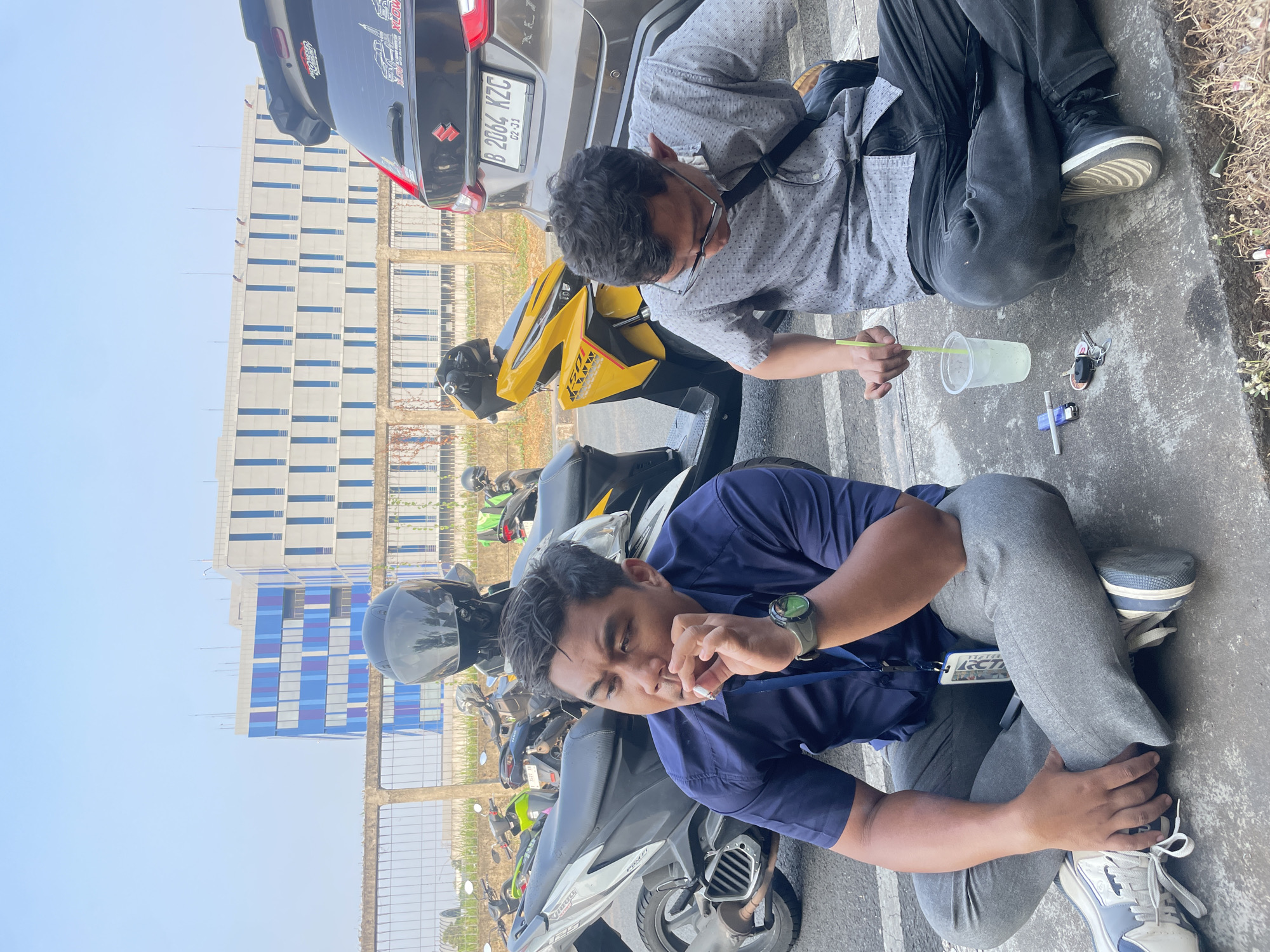 Two men are sitting on a paved surface, facing each other, with motorbikes and a car parked behind them. The man on the left is wearing a dark blue collared shirt and gray pants. He is holding a lit cigarette to his mouth, with ash visible. He has a watch on his left wrist. The man on the right is wearing a light gray patterned collared shirt and dark pants. He is holding a straw in a plastic cup filled with a light-colored liquid. Scattered on the ground near him are a cigarette lighter, keys, and an unlit cigarette. In the background, a modern building with blue and white panels is visible, along with a metal fence and parked motorbikes. The license plate of the car is visible and reads "B 2064 KZC". The scene appears to be outdoors, possibly in an urban or industrial area of Jatiwangi, Indonesia, given the presence of the building and vehicles. The sky is clear and blue.