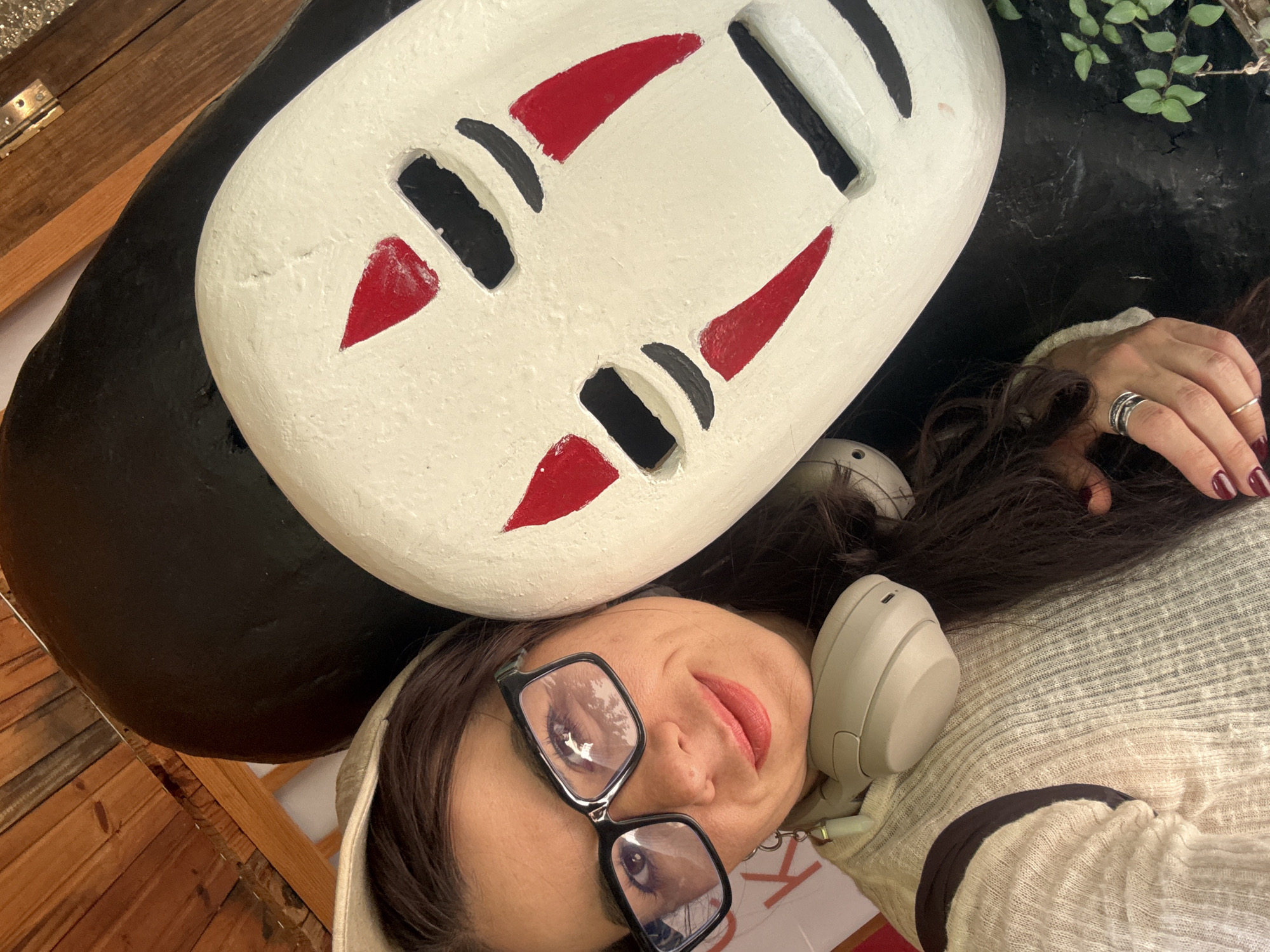 A woman is taking a selfie, leaning her head against a large, white mask with red and black markings. She is wearing a light-colored, textured sweater and glasses. Headphones are around her neck. The background appears to be an outdoor setting with wooden structures and some greenery. The location is identified as Lâm Viên - Đà Lạt, Vietnam.