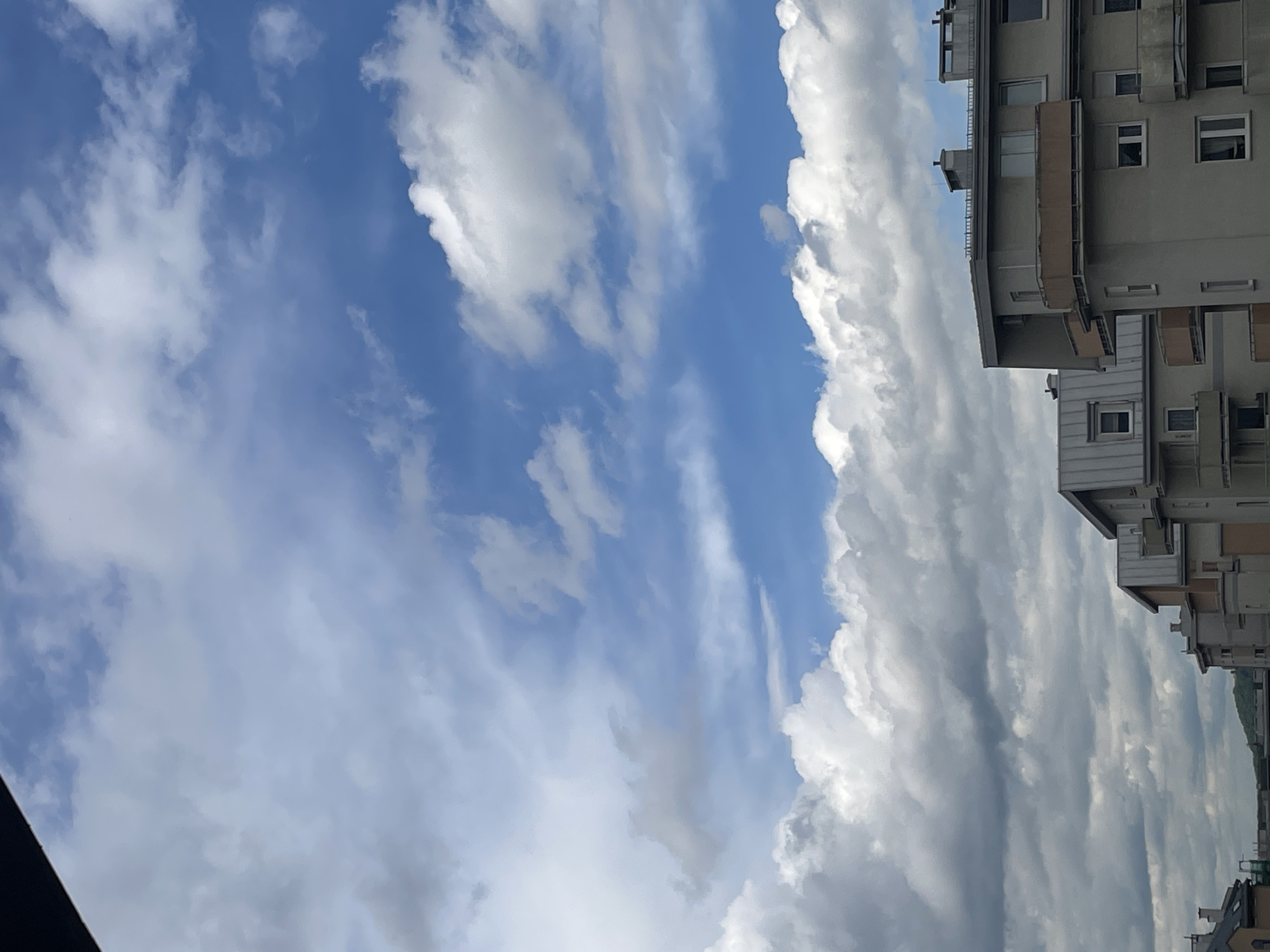 The image shows a view of the sky with clouds and the top portion of a building on the right side. The sky is predominantly blue with scattered white clouds of varying density. A section of a building with multiple stories is visible on the right side, featuring windows and balconies. The building appears to be made of gray concrete or a similar material. The image is taken from a low angle, looking upwards towards the sky and building. The location is Unknown, Unknown.