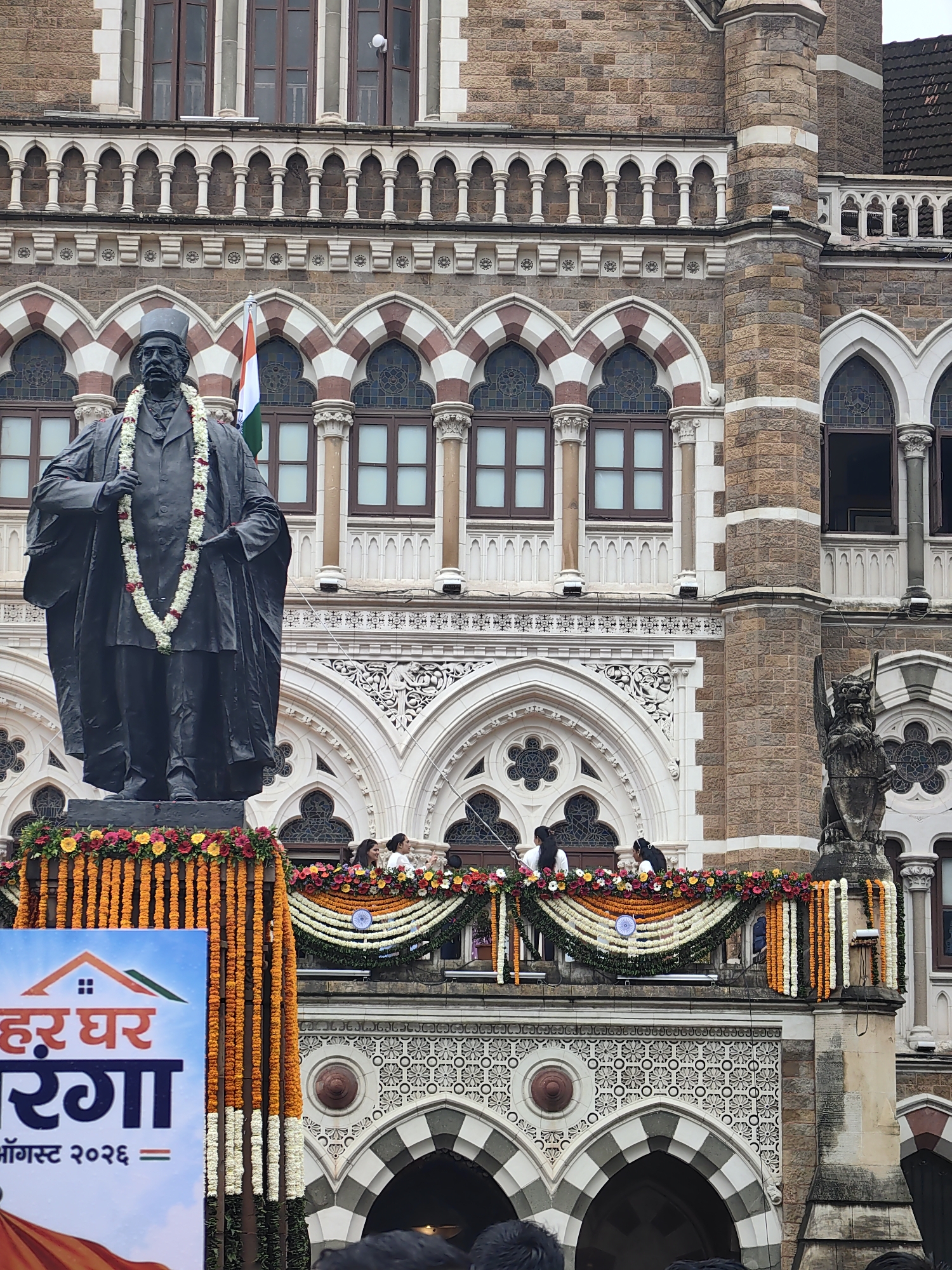 A statue of a man wearing a suit and a hat stands in front of a large stone building. The statue is adorned with a garland of flowers. A flag is visible behind the statue. People are standing on a balcony of the building, which is decorated with garlands of flowers. A banner with text and an image of a house is on the left side of the image. The building has arched windows and intricate architectural details. The scene appears to be set in a public space in Mumbai, India, possibly a government building or historical landmark.