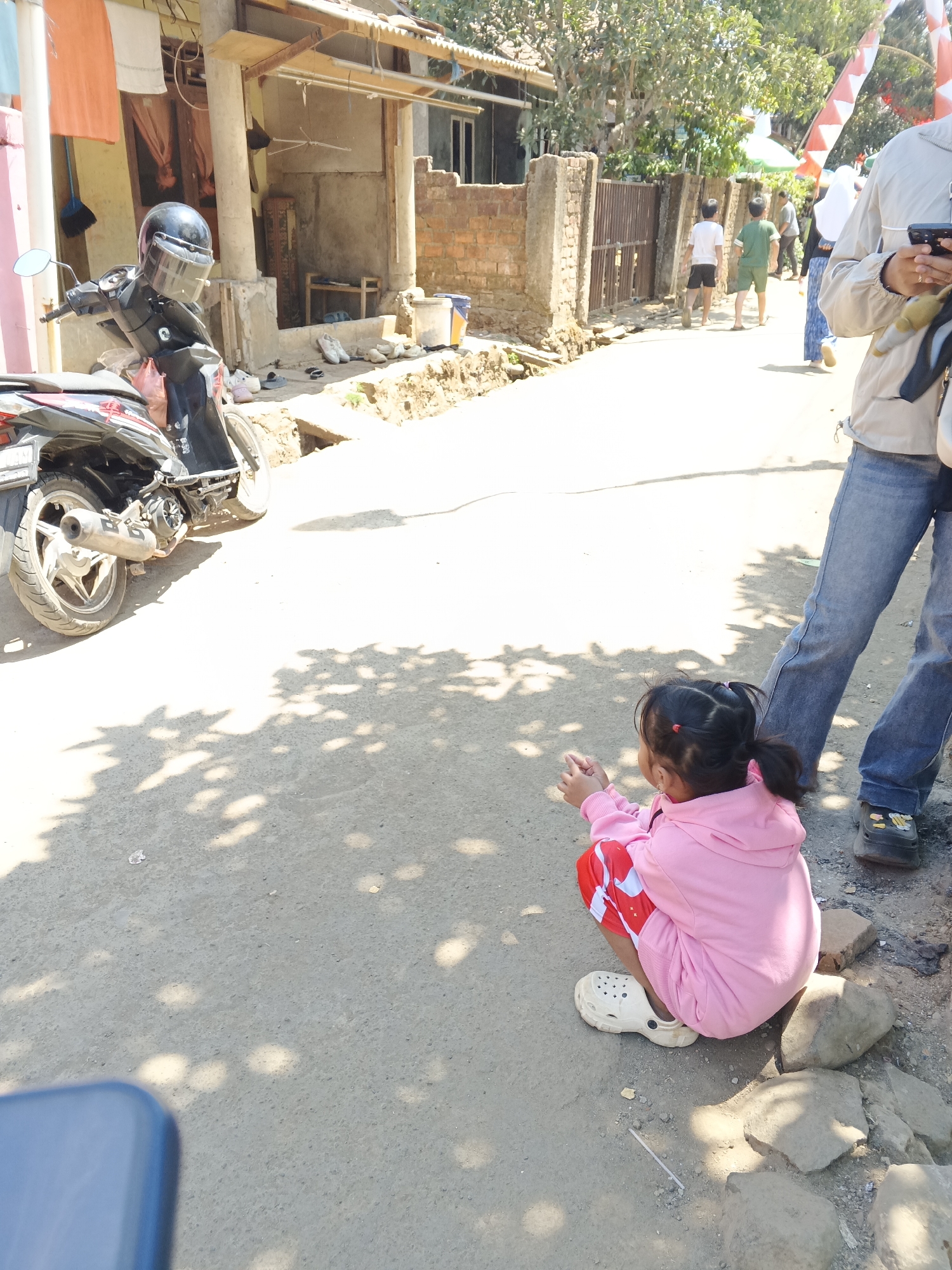 The image depicts an outdoor scene on a sunny day in Sukaharja, Indonesia. In the foreground, a young girl wearing a pink hoodie and red shorts with white patterns squats on the ground, facing away from the camera. She is wearing white clogs. To her right, the lower half of an adult's body, wearing blue jeans and brown shoes, is visible.
In the midground, a black scooter is parked on the left side of a paved road. It has a helmet with a clear visor on its handlebars. Behind the scooter, a low-slung building with a tiled roof and brick walls is visible. Laundry hangs from a line in front of the building. Further down the road, several people, including children and adults, are walking. Some are wearing casual clothing, and there are red and white Indonesian flags decorating the area.
The environment is characterized by a dusty, unpaved roadside with scattered rocks and debris. Sunlight casts shadows of trees onto the ground, suggesting a dappled shade. The overall impression is a casual street scene in a residential area.