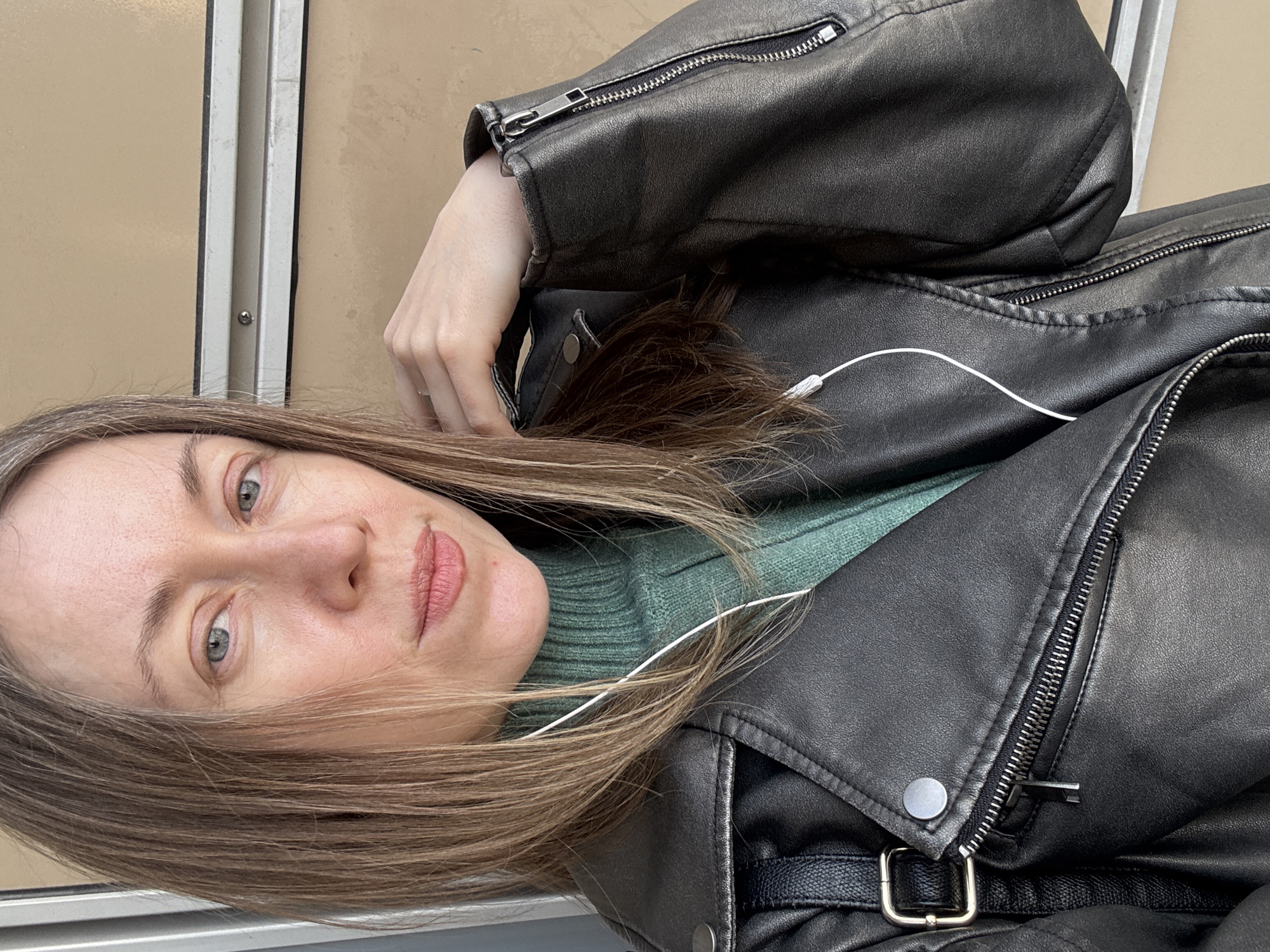A woman with light brown hair and grey eyes is wearing a black leather jacket over a green turtleneck sweater. She has white earbuds plugged into her ears. The background features a beige wall with vertical silver trim. The location is Saint Petersburg, Russia.