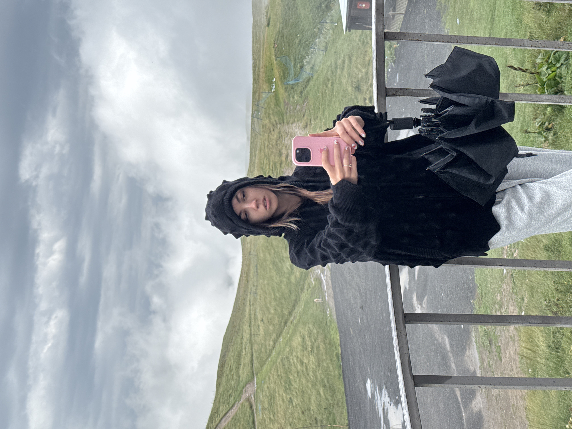 In Gudauri, Georgia, a woman stands on a balcony, holding a pink smartphone to take a selfie. She is wearing a black hooded sweatshirt with a textured design and light gray sweatpants. An open black umbrella rests on the railing in front of her. The background features a grassy hillside with a winding path, indicative of a mountainous or ski resort area, under a cloudy, overcast sky.